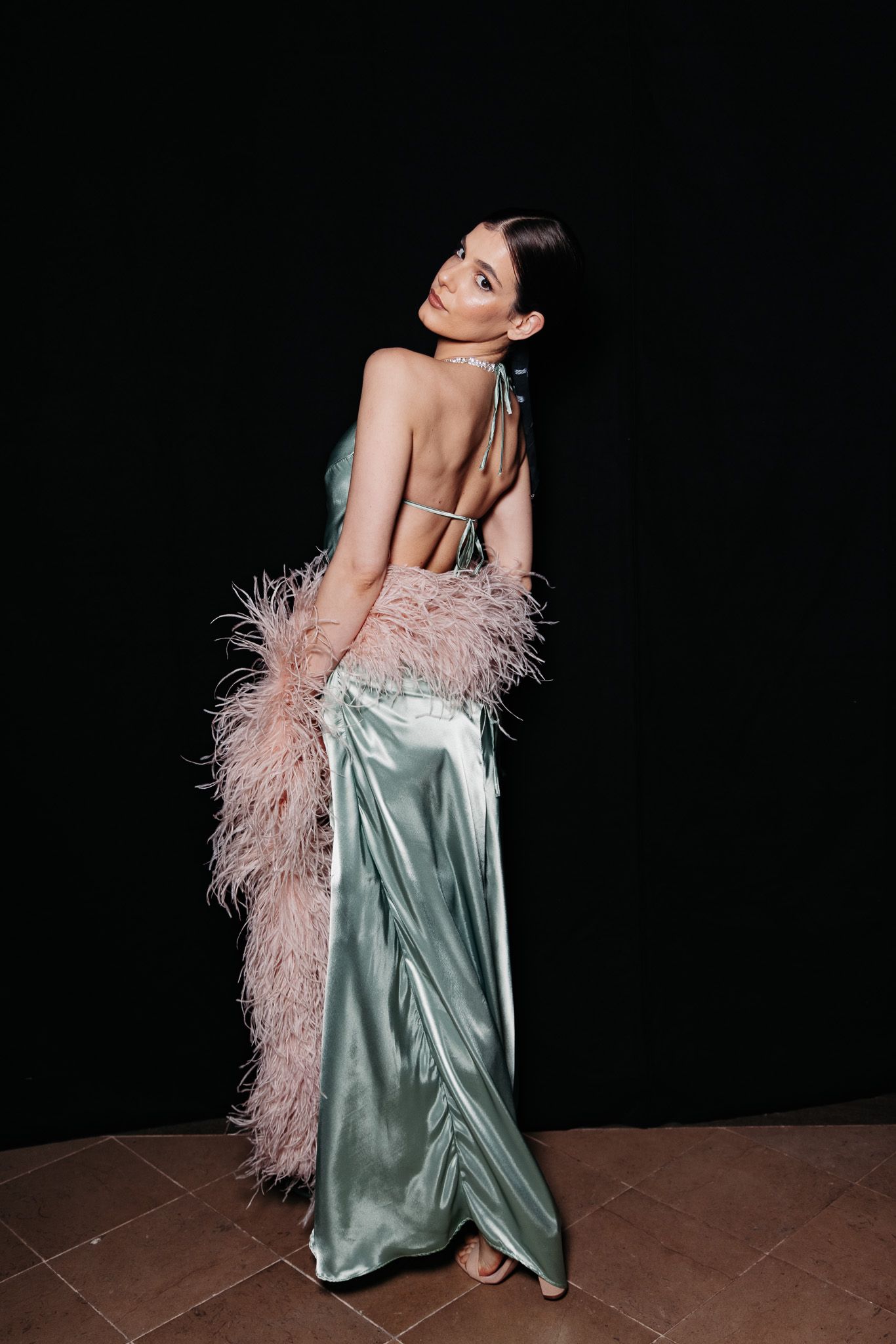 Model poses in a glamorous silver satin gown with pink feather boa against dark background in fashion photoshoot series.