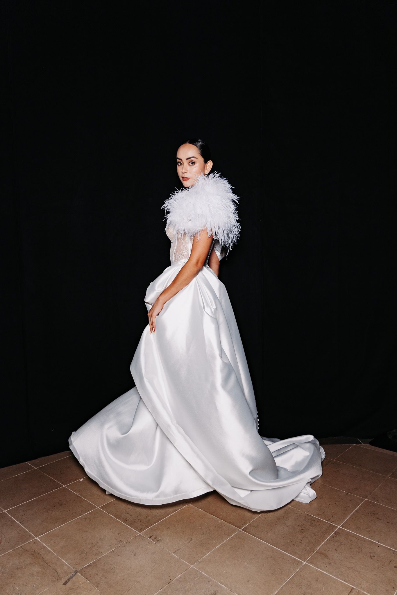 Elegant white wedding gown with dramatic feather collar and flowing train photographed against dark background.
