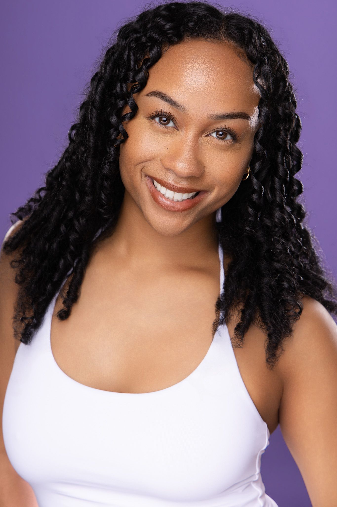Smiling young woman with long curly braids wearing a white tank top against a purple background.