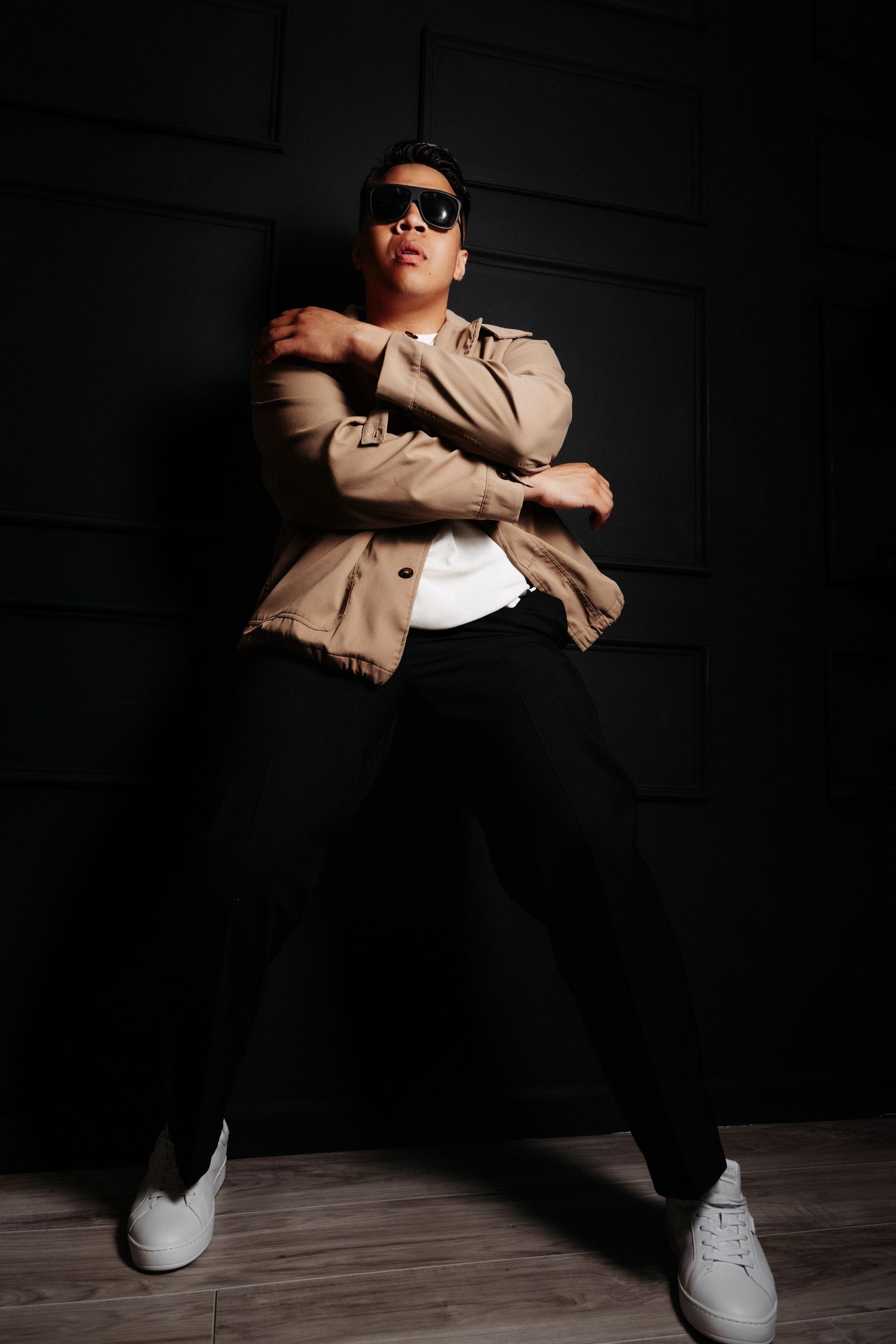 Person in beige jacket and dark pants sits in dramatic lighting with white sneakers against dark background.