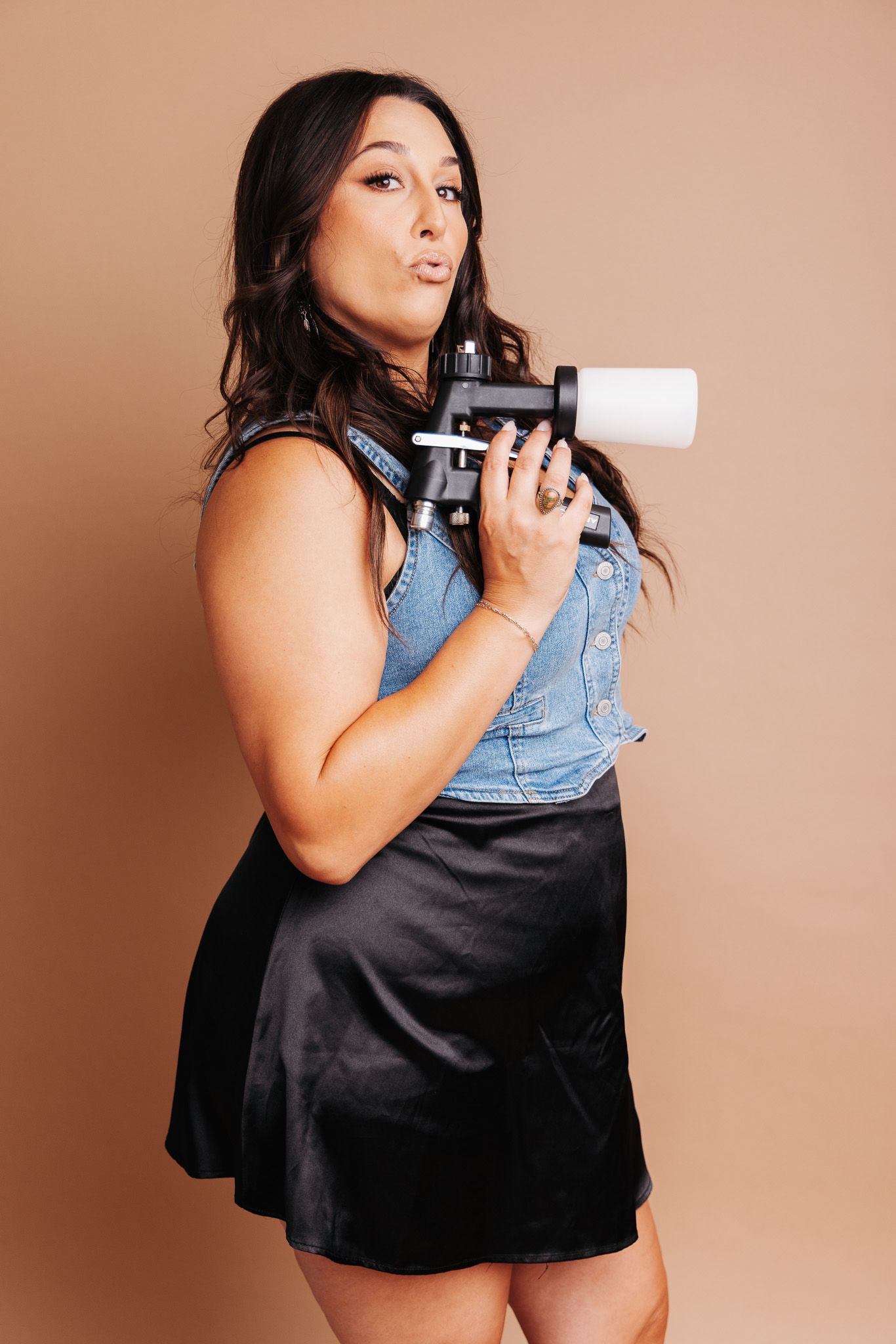 A woman in a denim vest and black dress poses with playing cards against a warm brown background.