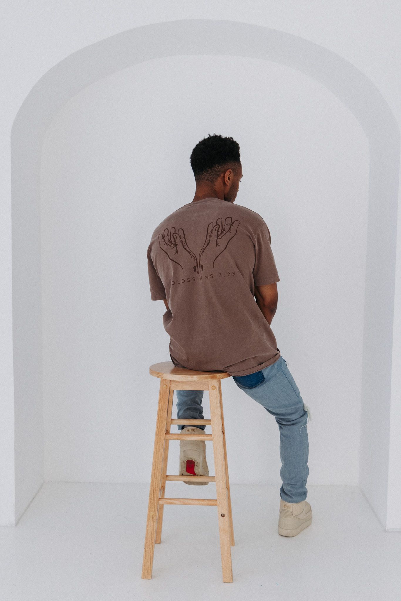 Back view of person wearing brown t-shirt and jeans sitting on wooden stool in white archway.