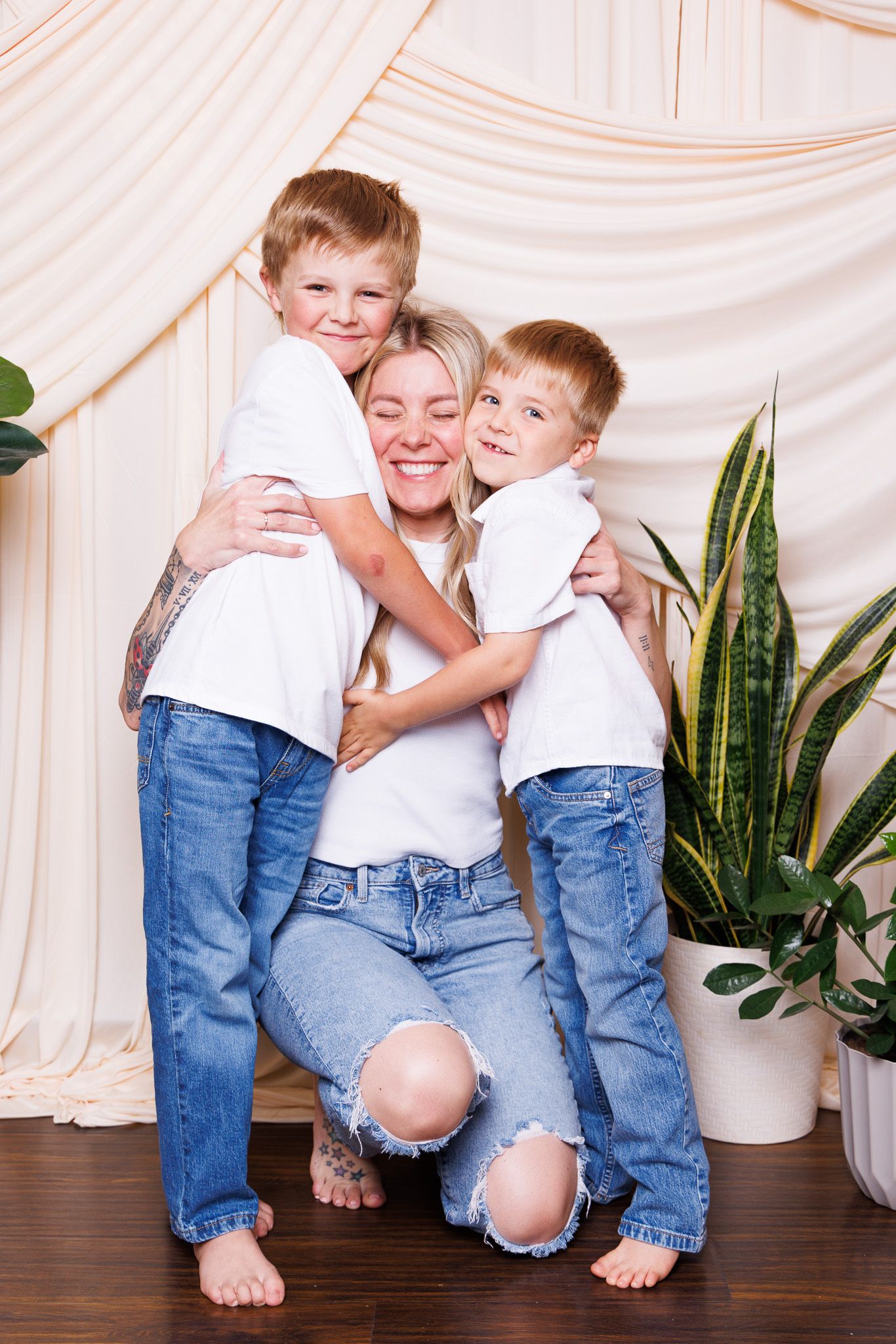 A smiling mother in white tee and ripped jeans hugs her two blonde sons, all barefoot, posed before draped fabric backdrop.