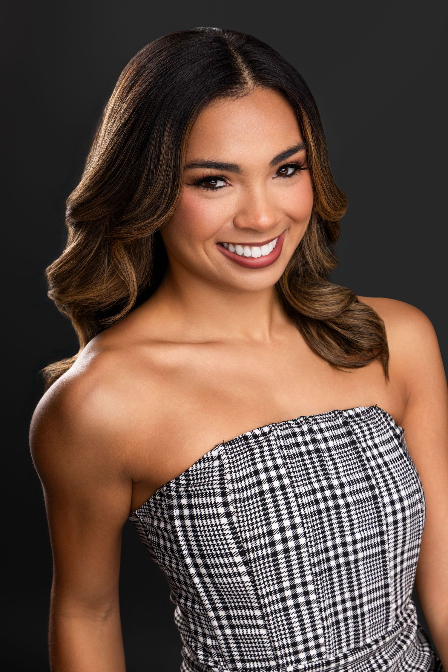 Portrait of a smiling person with wavy brown hair wearing a strapless black and white plaid top against a dark background.