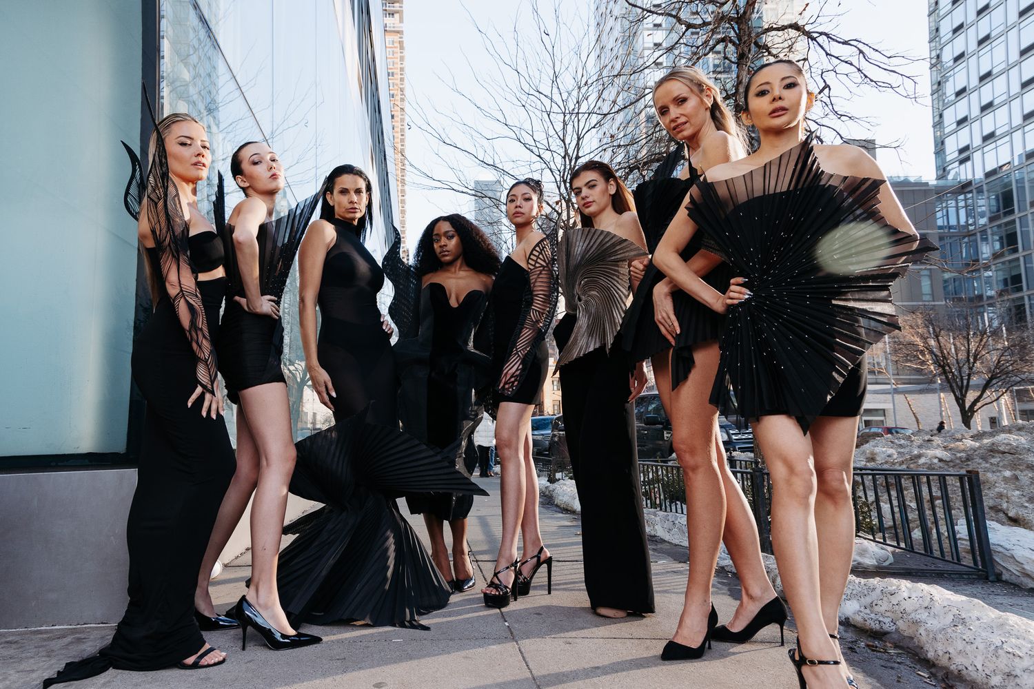 Group wearing black dresses poses outdoors in urban setting for fashion shoot.