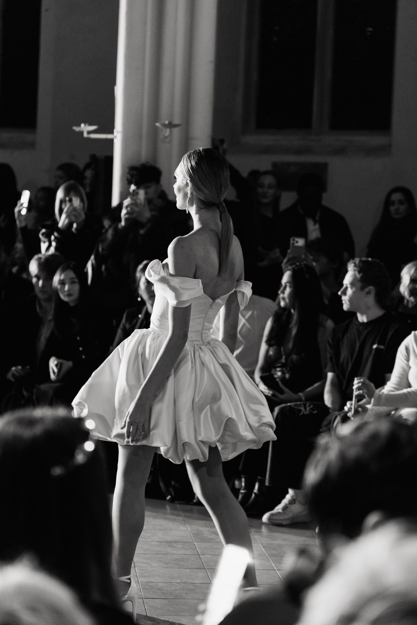 Black and white photo of a fashion show with models walking the runway in front of a seated audience.