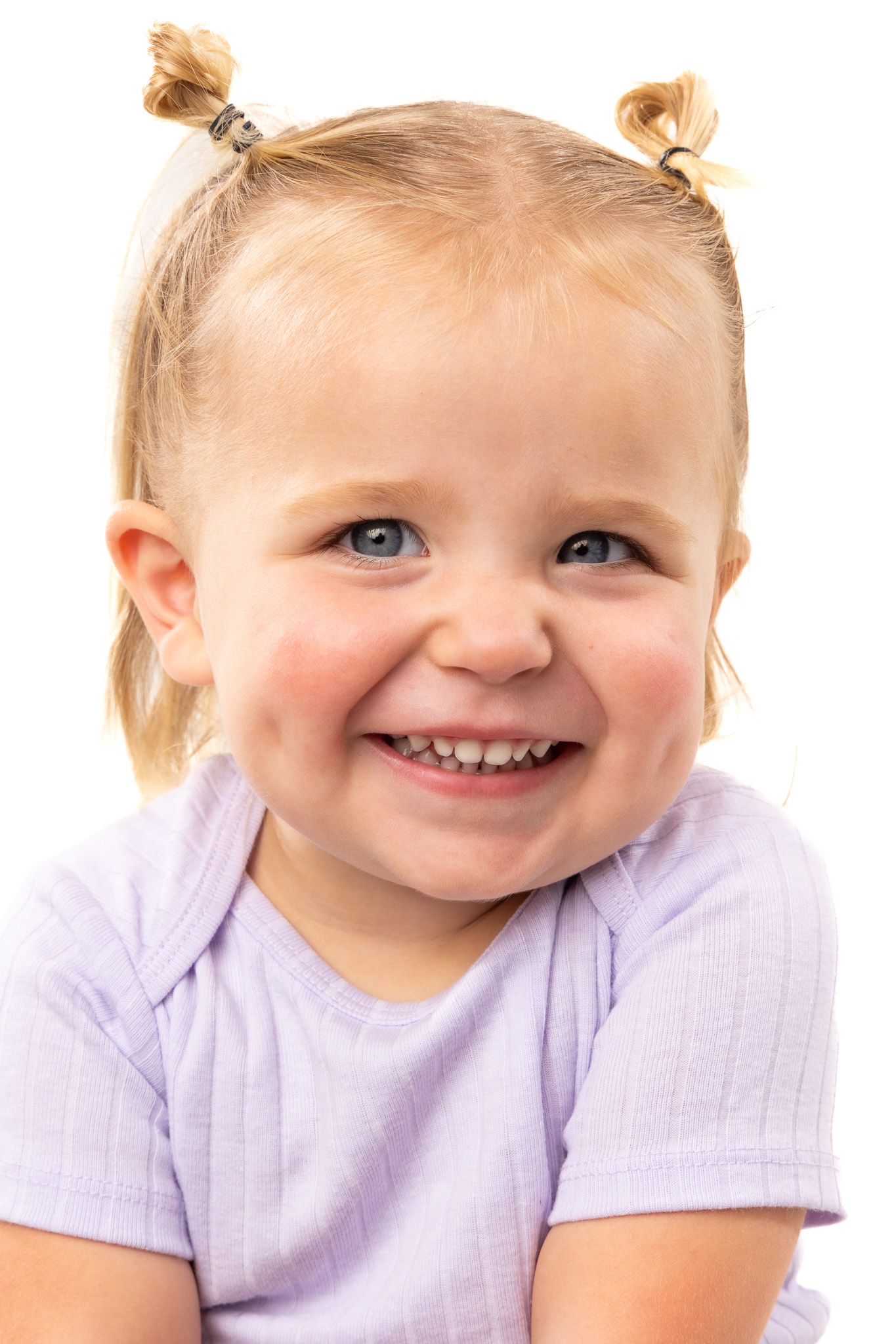 Young child wearing a purple shirt smiles brightly with blonde hair styled in two small buns.