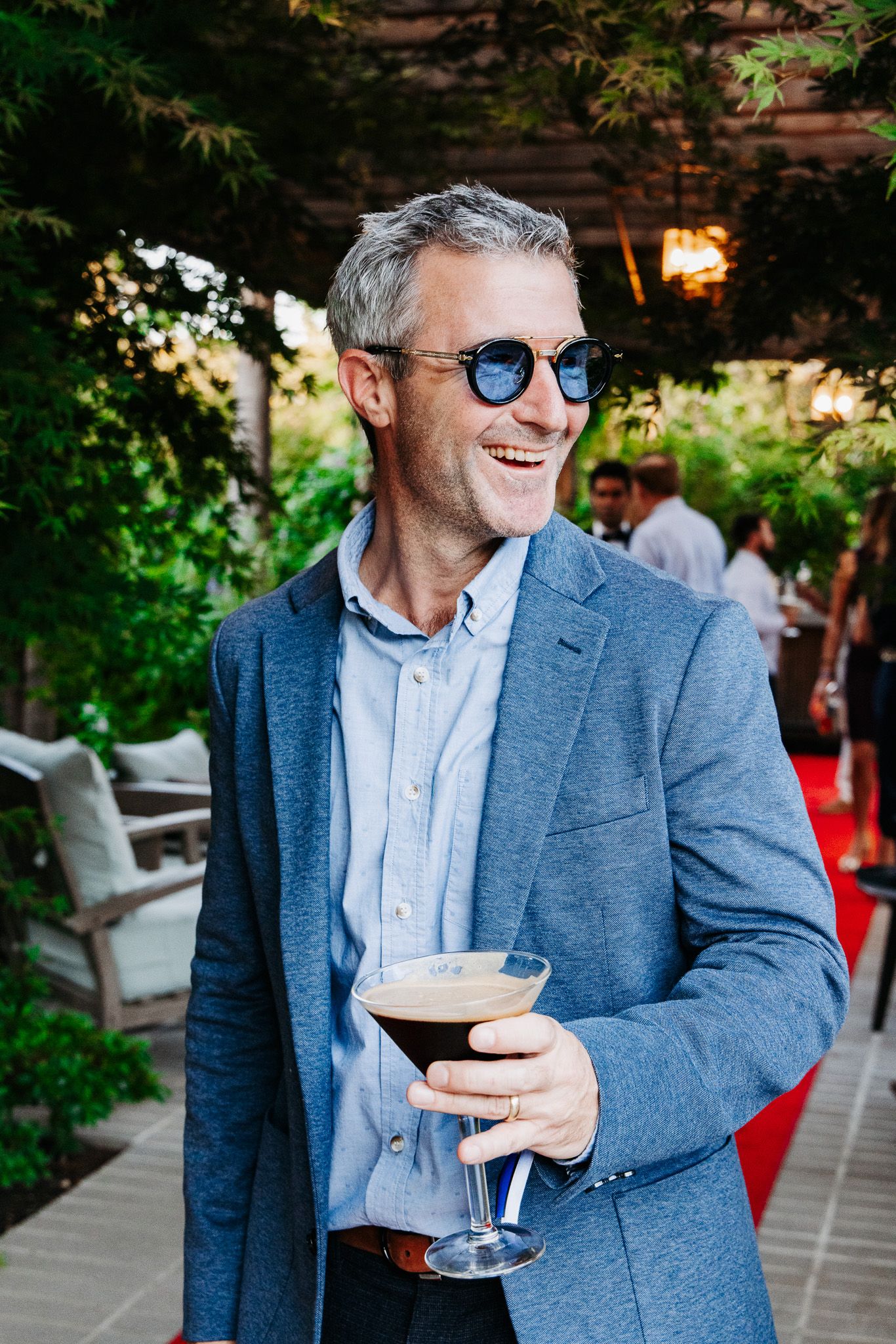 Stylish person in blue blazer and sunglasses enjoying cocktails at an outdoor venue with string lights.