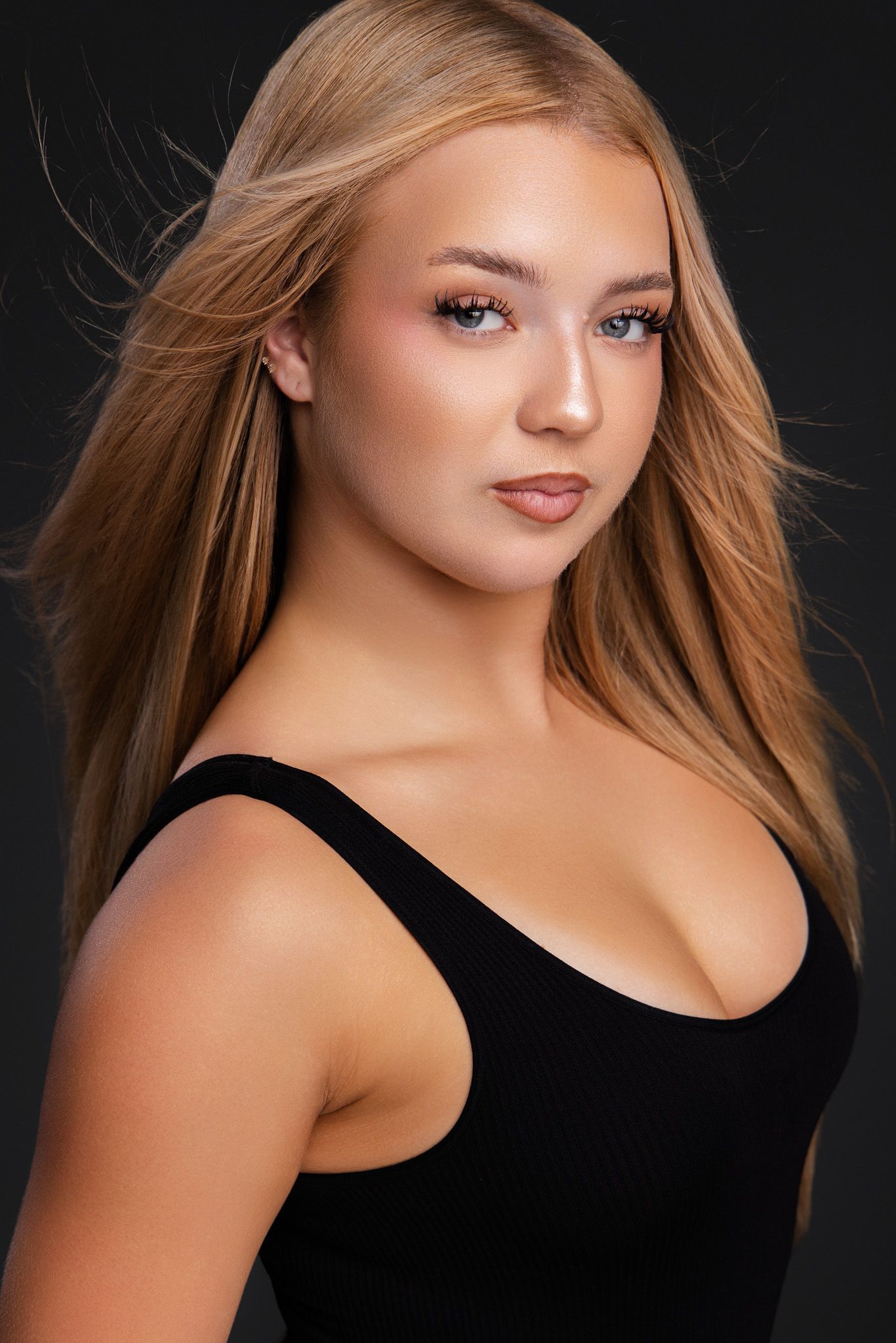 Young blonde woman in black tank top poses against dark background with flowing hair and natural makeup.