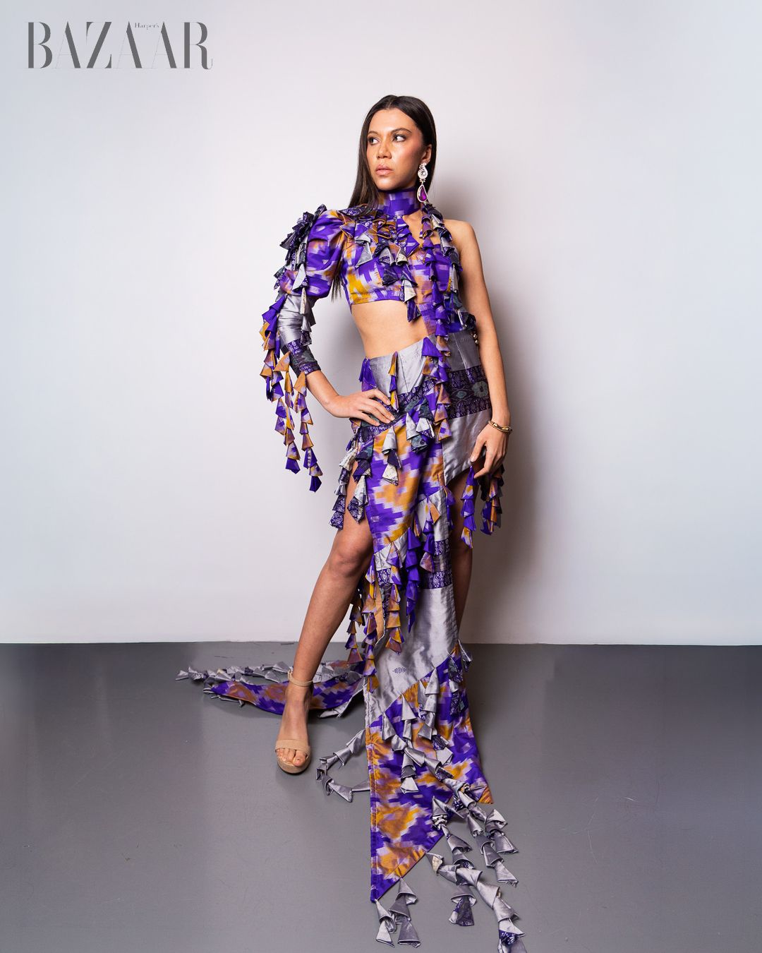 Model poses in Harper's BAZAAR wearing a purple floral cutout gown with cascading fabric details and fringe accents.