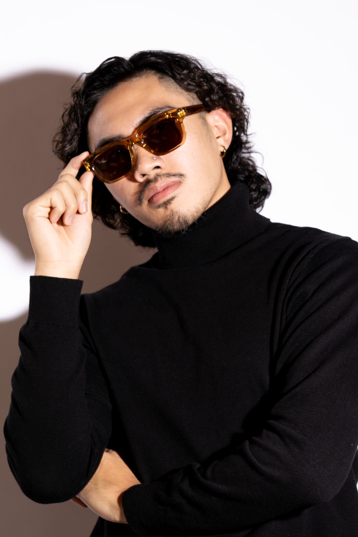 Stylish man in black turtleneck adjusting tortoiseshell sunglasses, with curly hair and small beard.