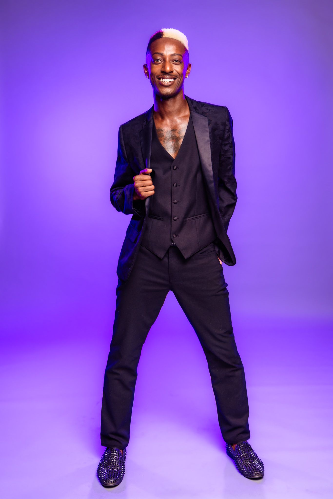Stylish person in black suit and vest poses confidently against purple backdrop.