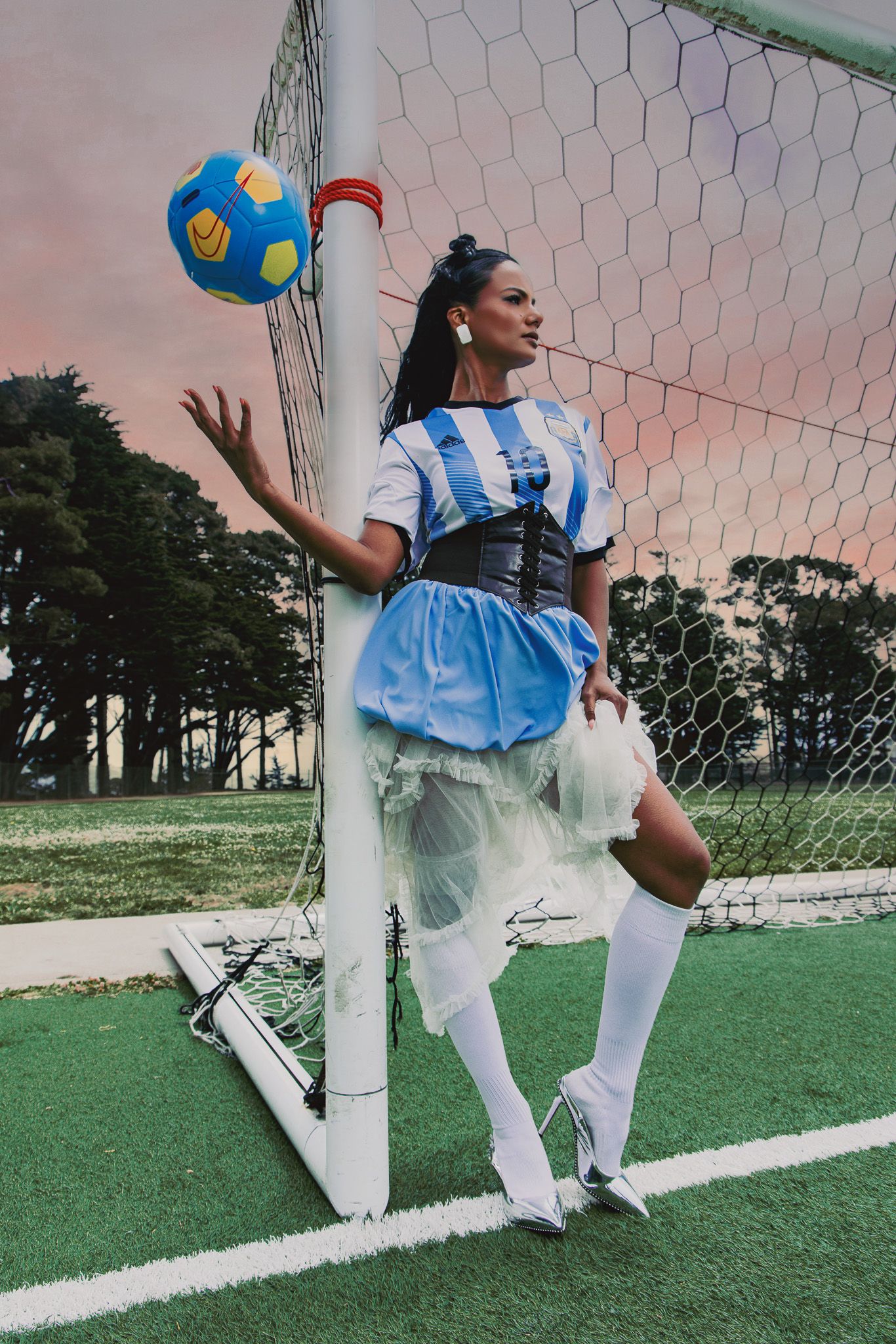 Woman in Argentine soccer jersey and tutu skirt poses with ball at goal post on green field at dusk.
