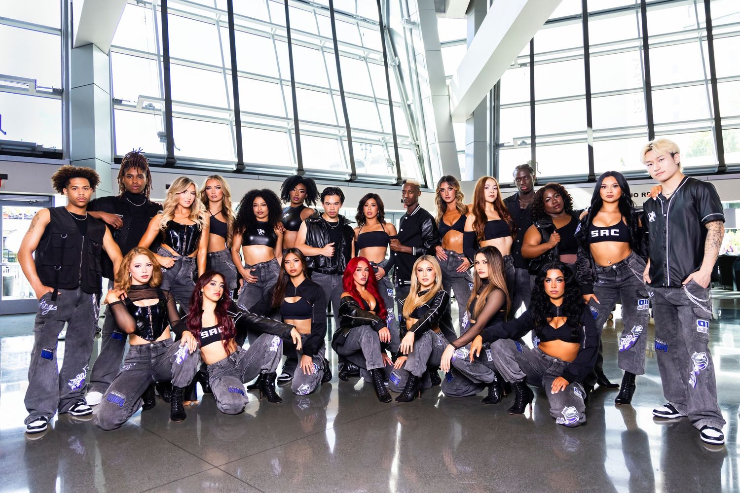 Large diverse dance crew posing together in matching black outfits and cargo pants inside a modern building.