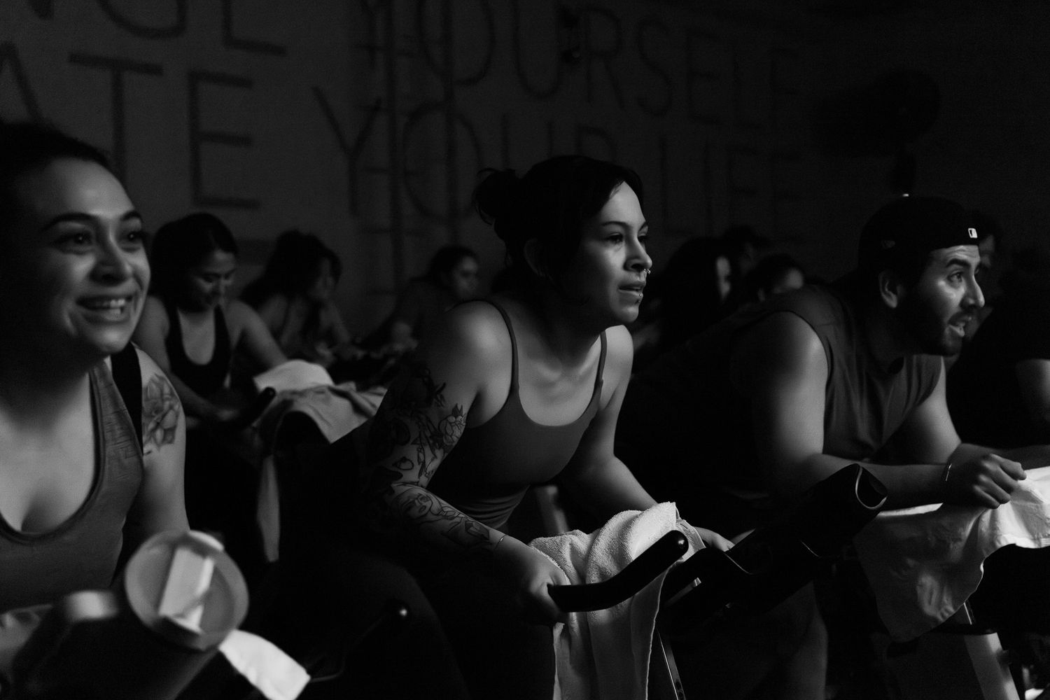 Indoor cycling class participants riding exercise bikes in a dark studio setting.