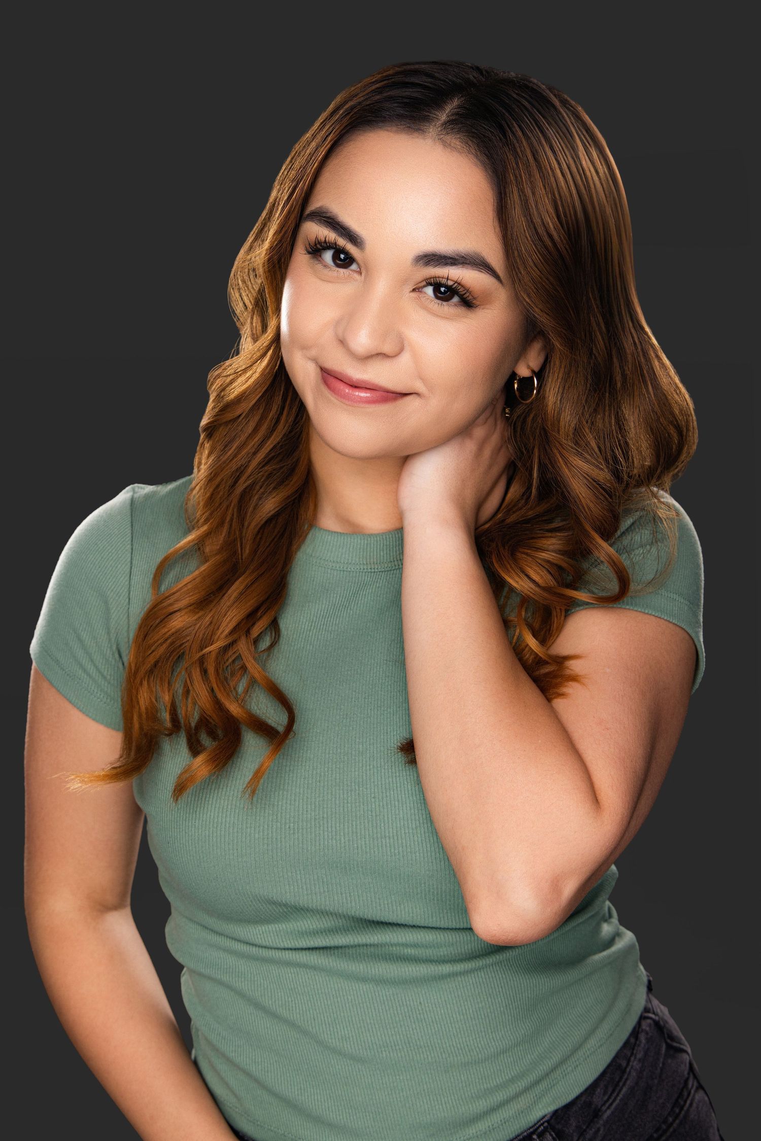 Portrait of a young woman with wavy brown hair wearing a sage green t-shirt against a dark background.