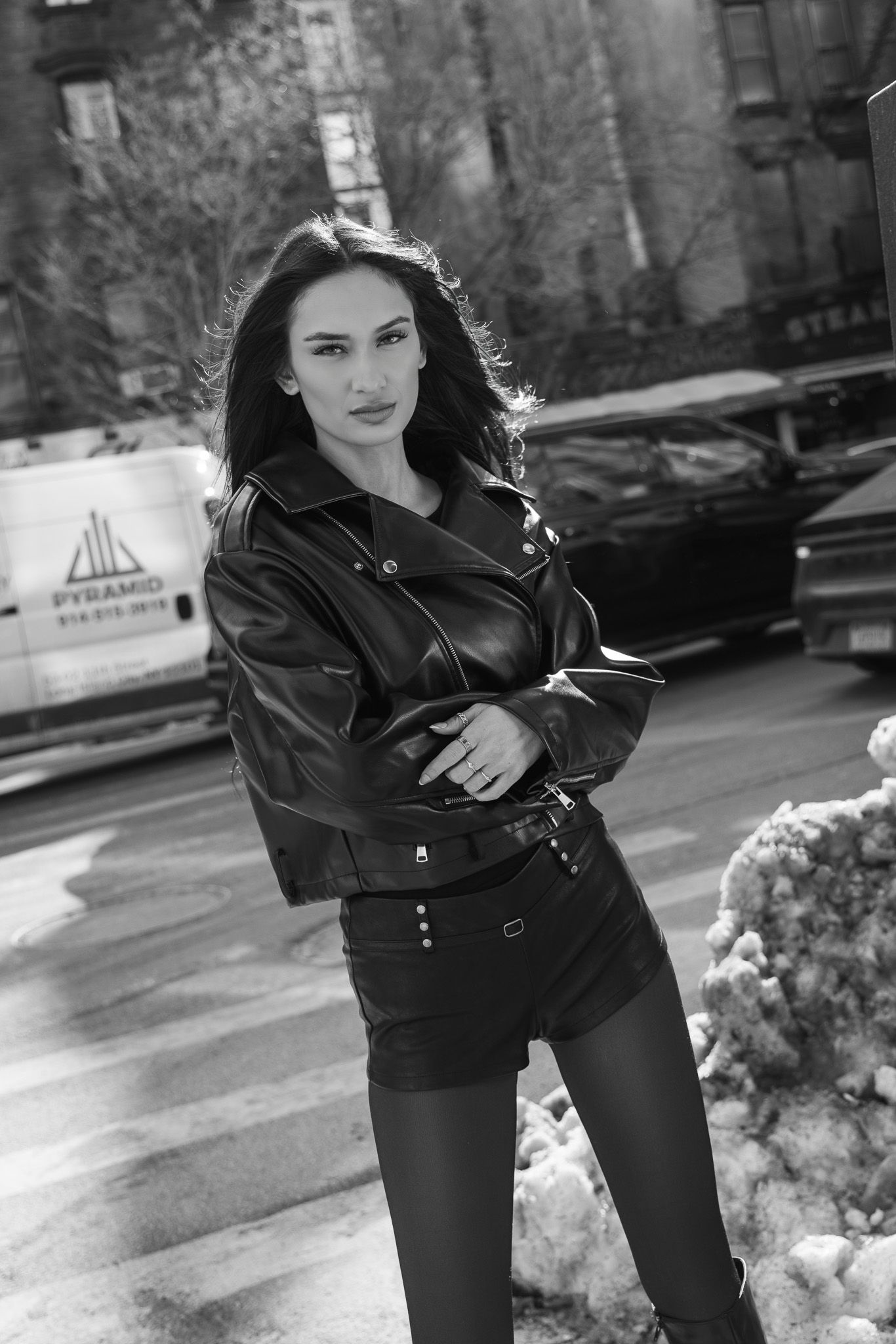 Black and white portrait of someone in an edgy leather jacket and shorts on a city street with snow in background.