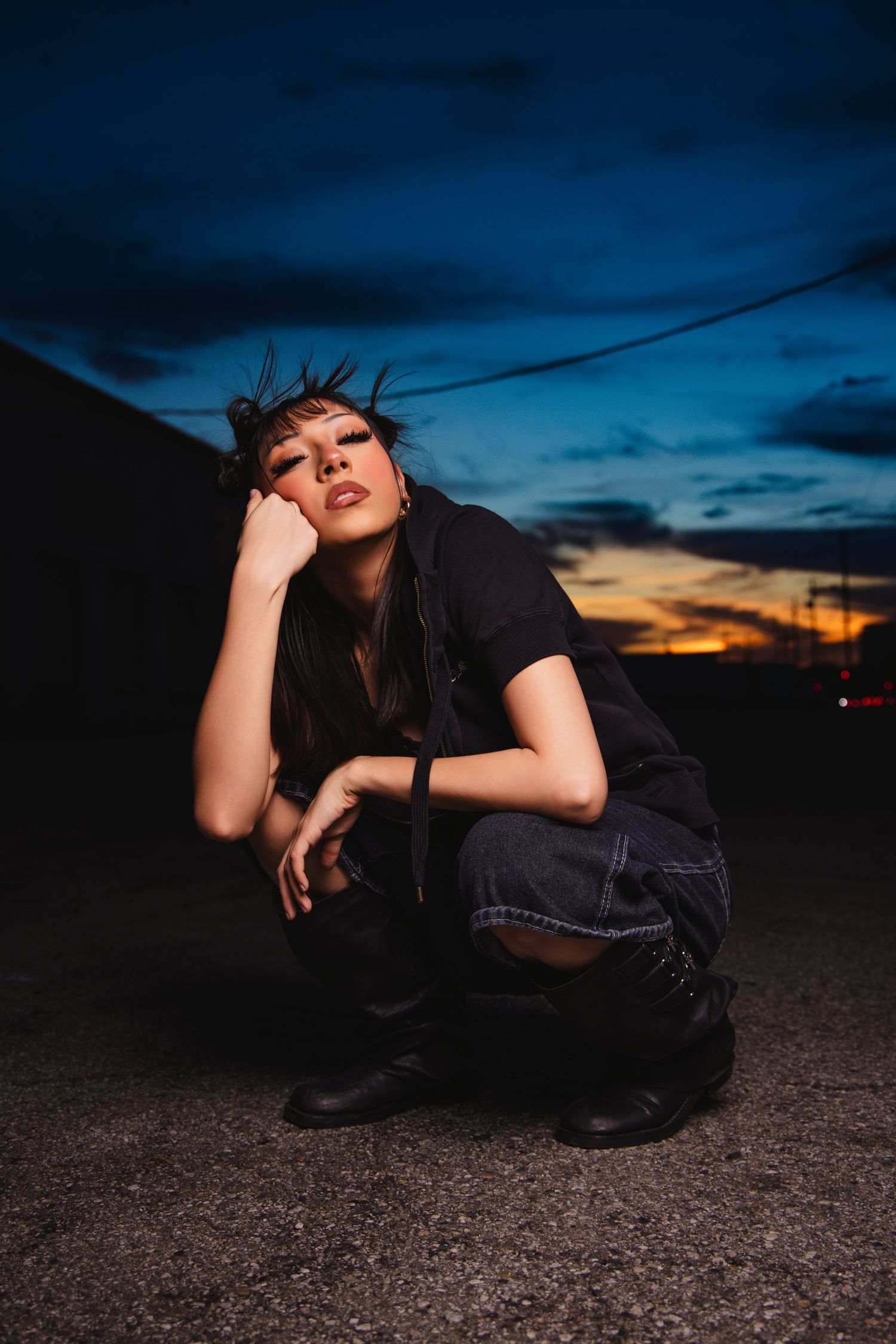 A person in dark clothing poses dramatically on the ground against a dusky blue and orange sunset sky.
