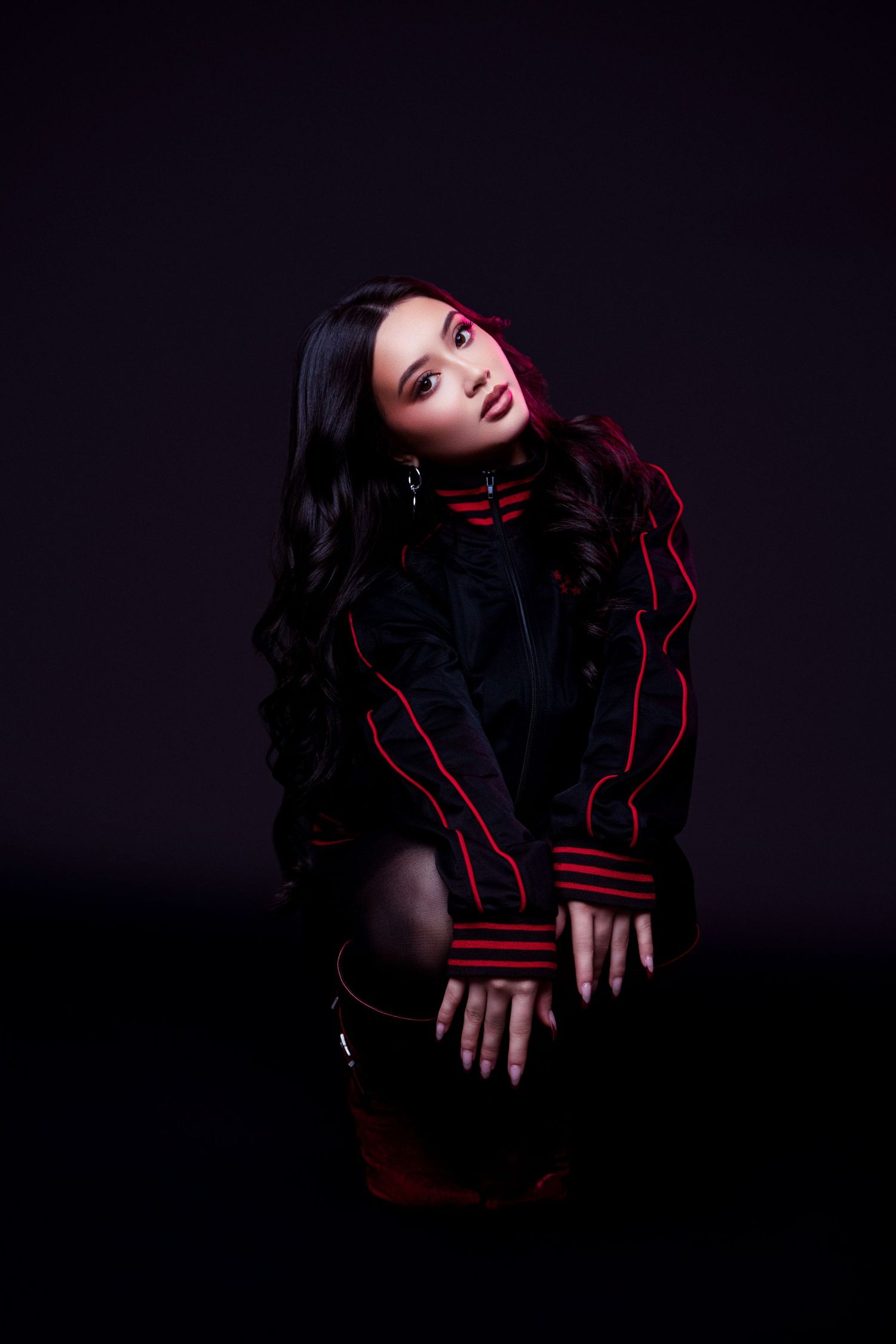 Dramatic fashion portrait in dark setting with red accent lighting showing model in black outfit with red trim details.