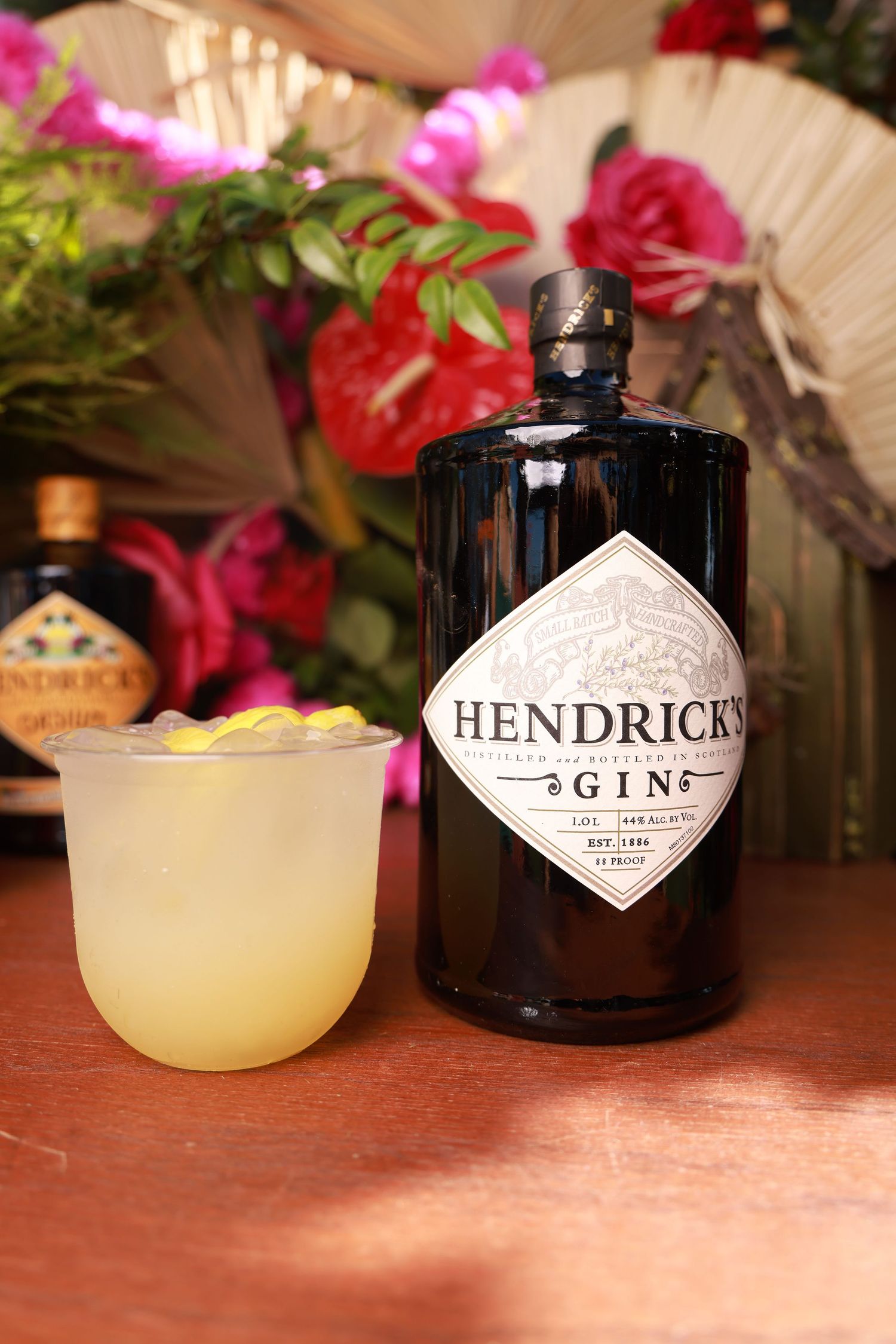 A yellow cocktail served in a coupe glass next to a Hendrick's gin bottle with pink flowers in the background.