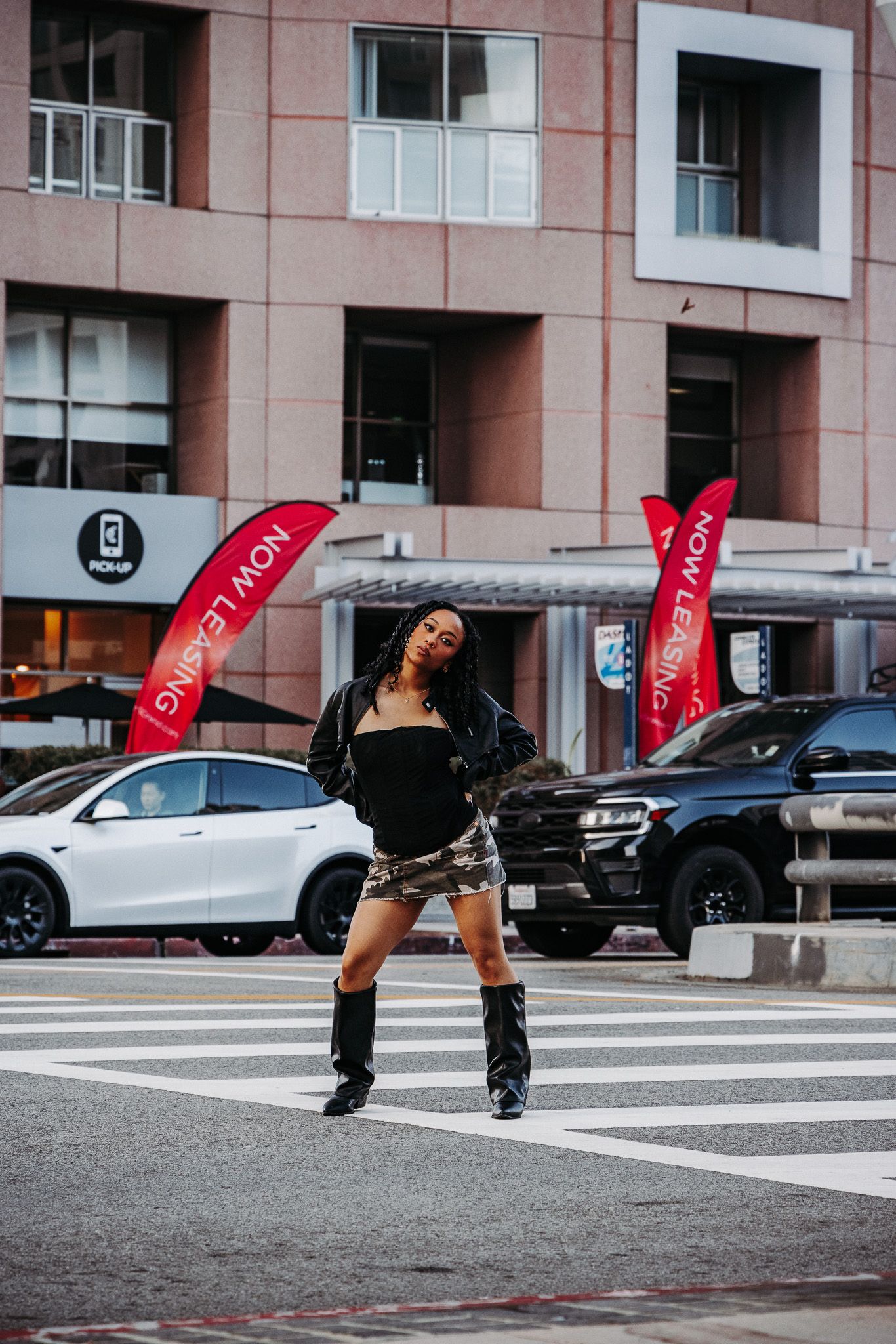 A fashionable person poses energetically on a city crosswalk with red banners and parked cars in the background.