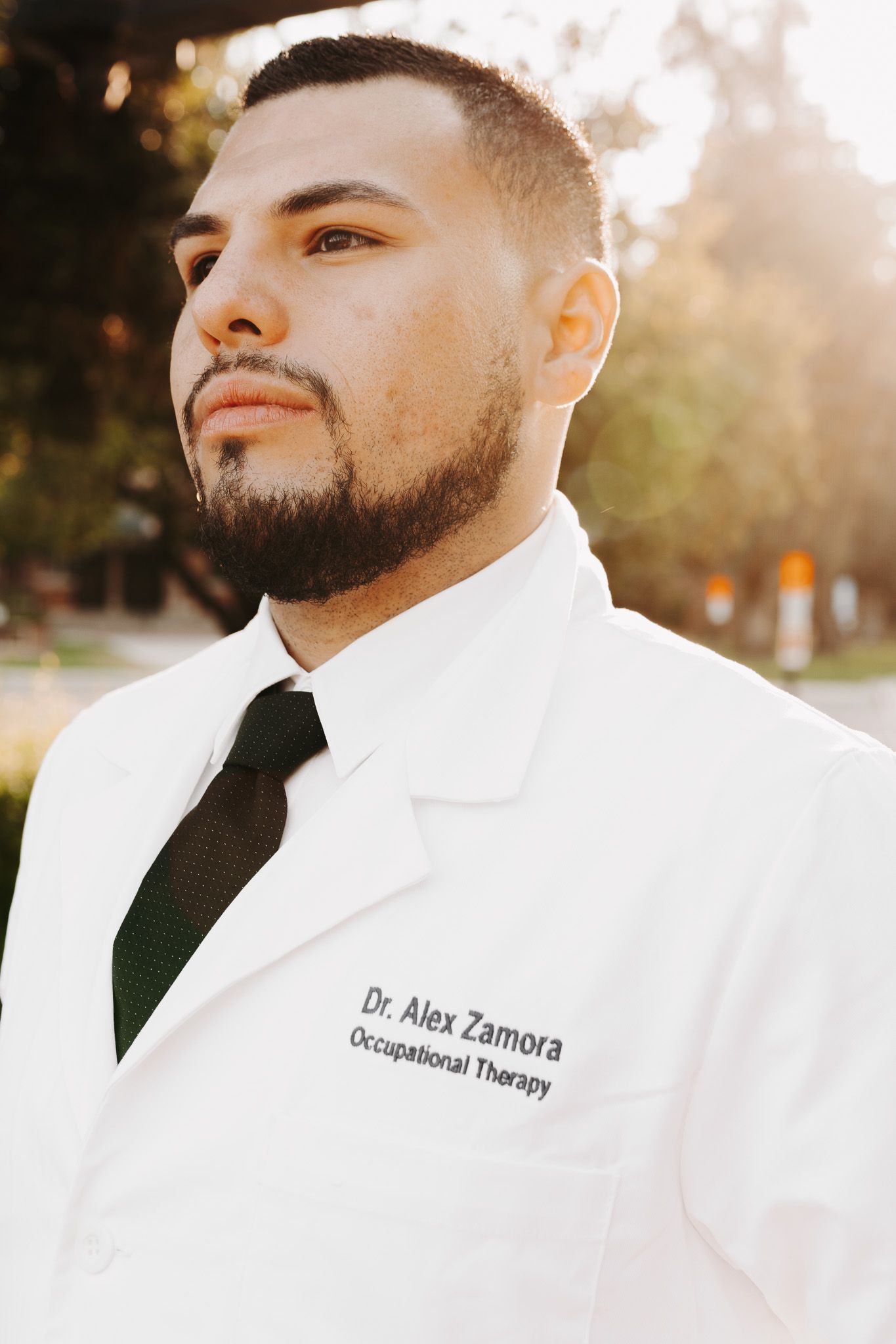 Healthcare professional wearing a white medical coat and black tie smiles in outdoor sunlit setting.