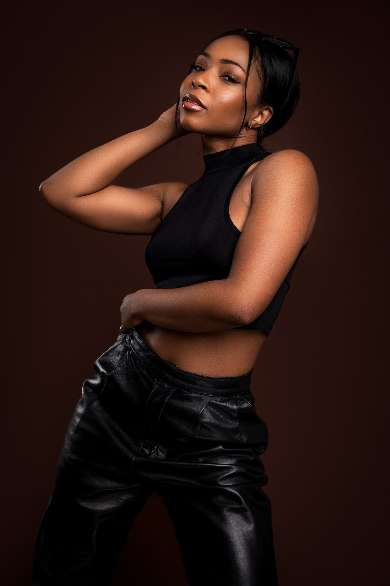 Artistic fashion portrait with black crop top and leather pants against brown background in dramatic lighting.