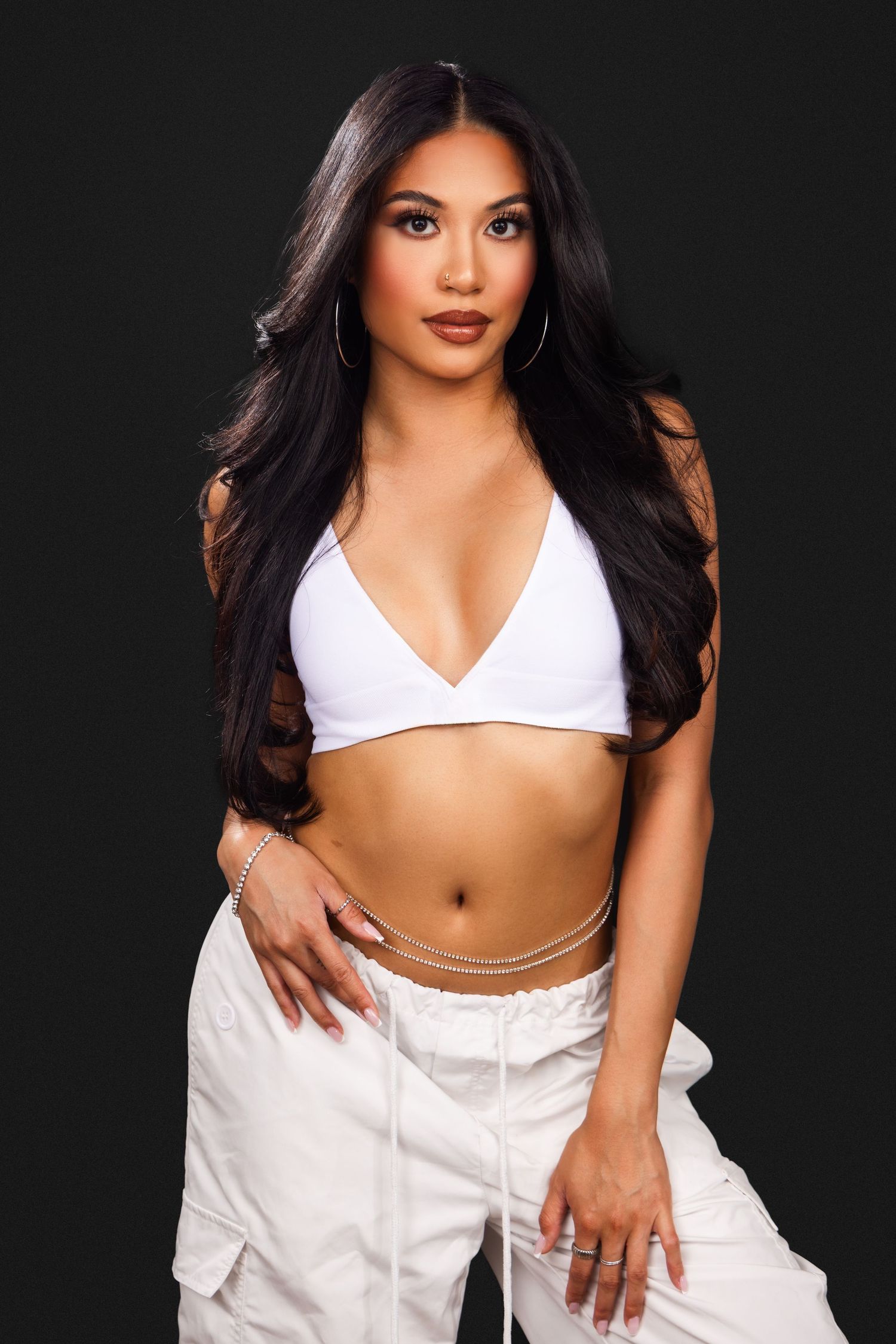 Fashion portrait in white crop top and cargo pants with long dark hair against black background.