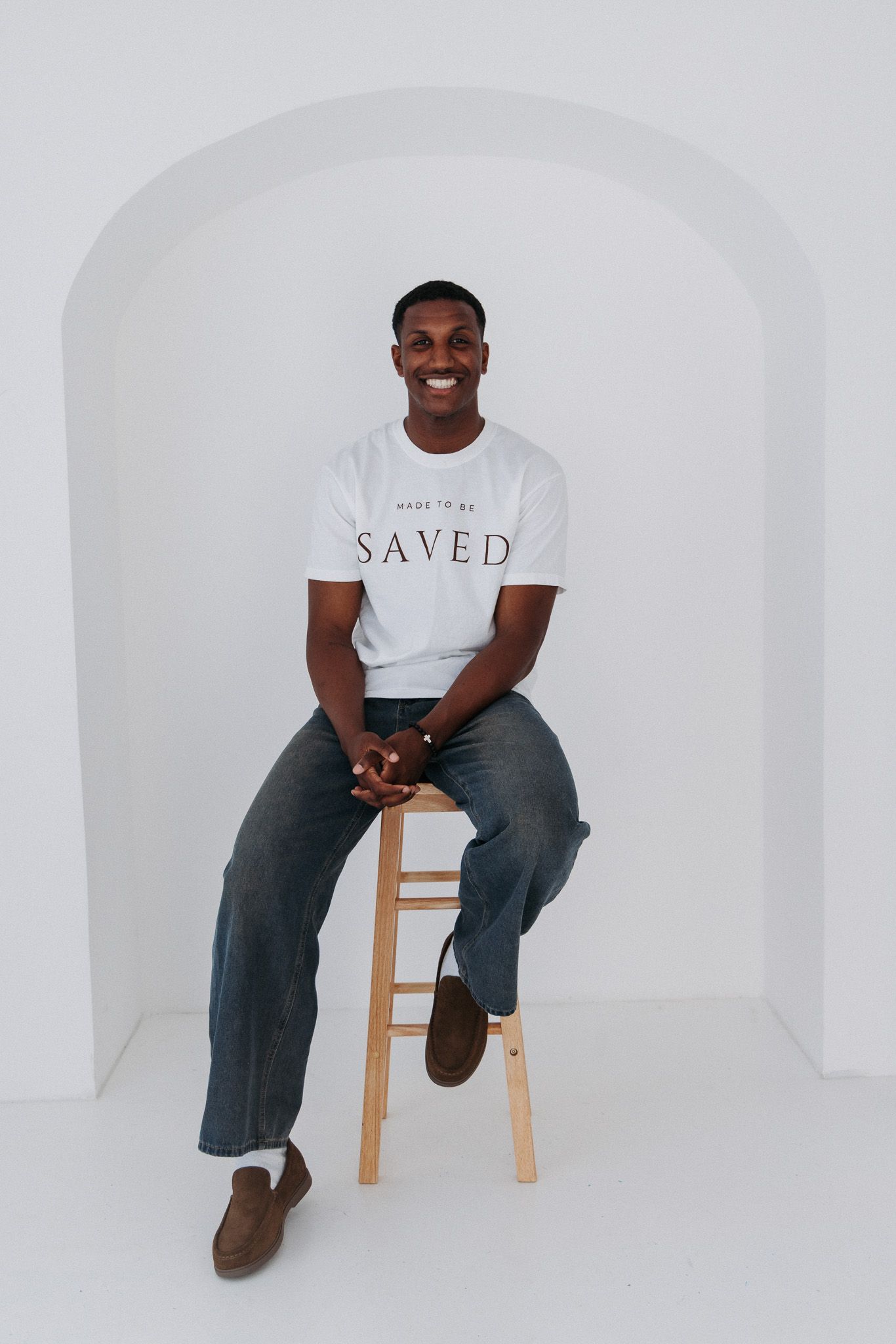 Model seated on wooden chair wearing white t-shirt and jeans in clean white studio setting.