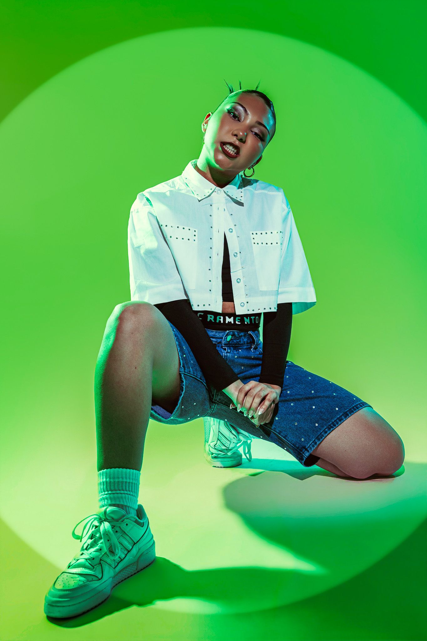 A stylish woman poses in a white crop top, denim skirt, and green sneakers against a vibrant green background.