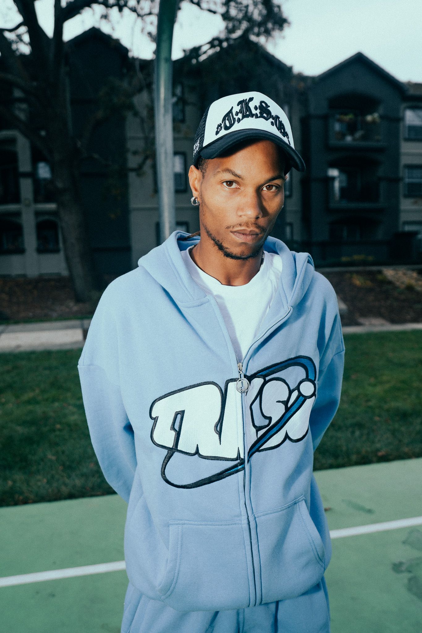 Young man in a light blue graphic zip hoodie and black cap poses on an outdoor basketball court.
