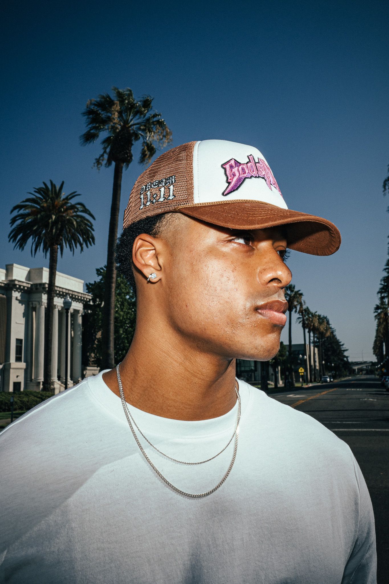 Young man wearing a brown and white trucker hat, silver chain necklace, and white t-shirt with palm trees behind him.