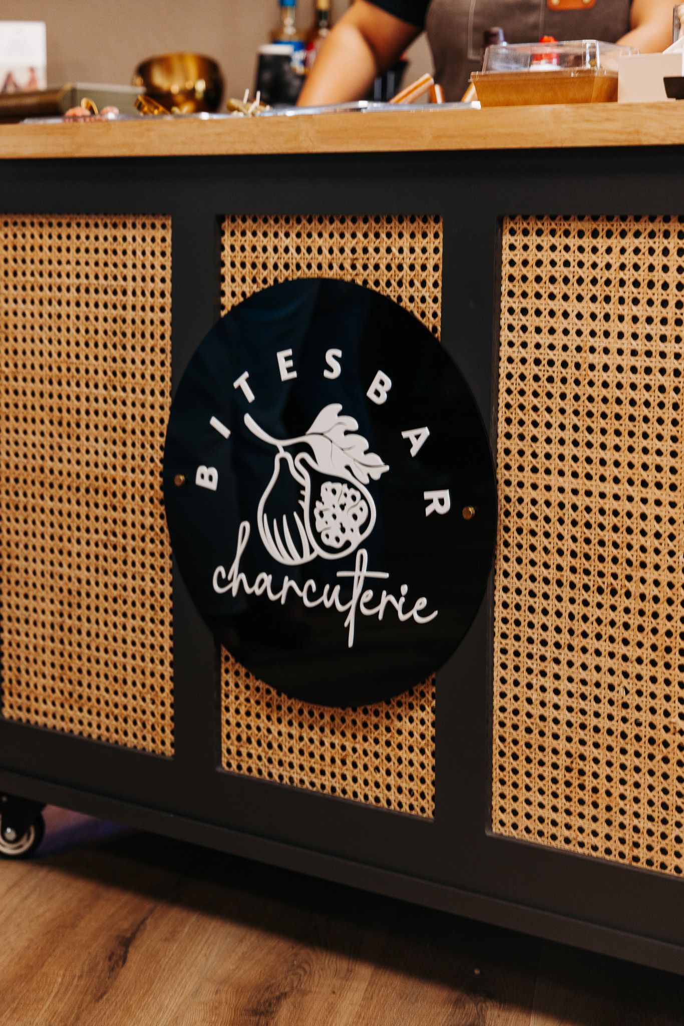 Black circular Bites Bar Charcuterie sign on a rattan cane panel counter at a food vendor booth.