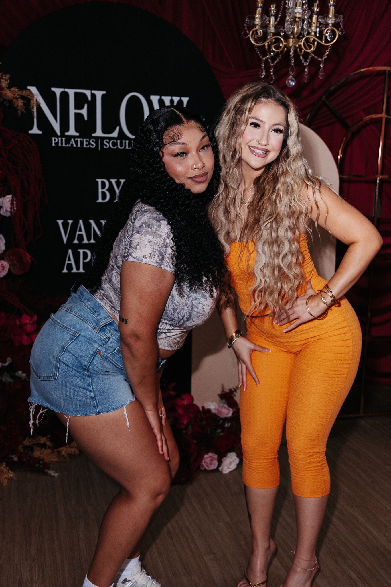 Two women pose together at a nightclub event with NFLOW signage and chandelier in background.