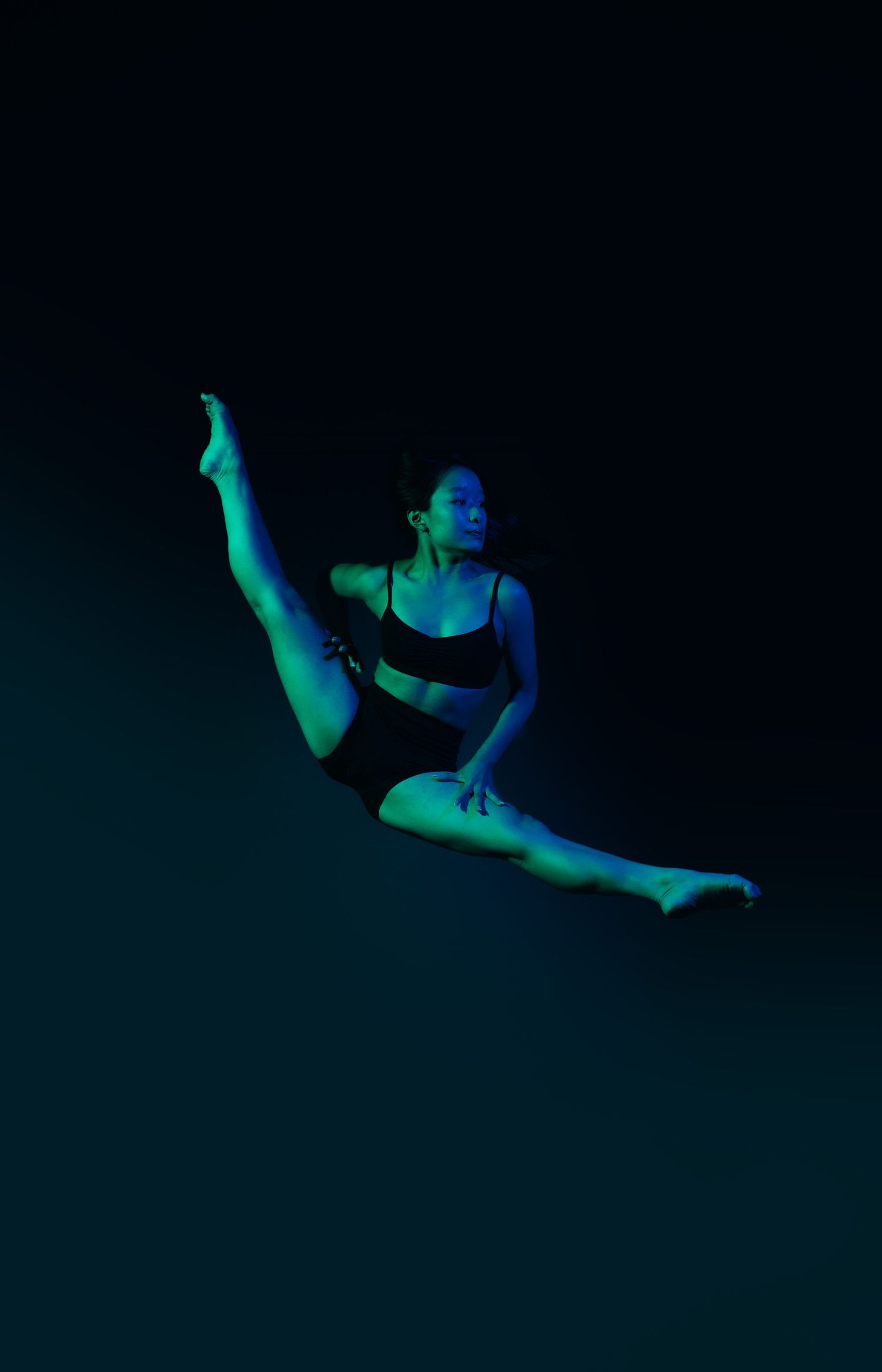 A dancer performs expressive movements in a dark setting with ethereal blue-green lighting creating a mystical atmosphere.