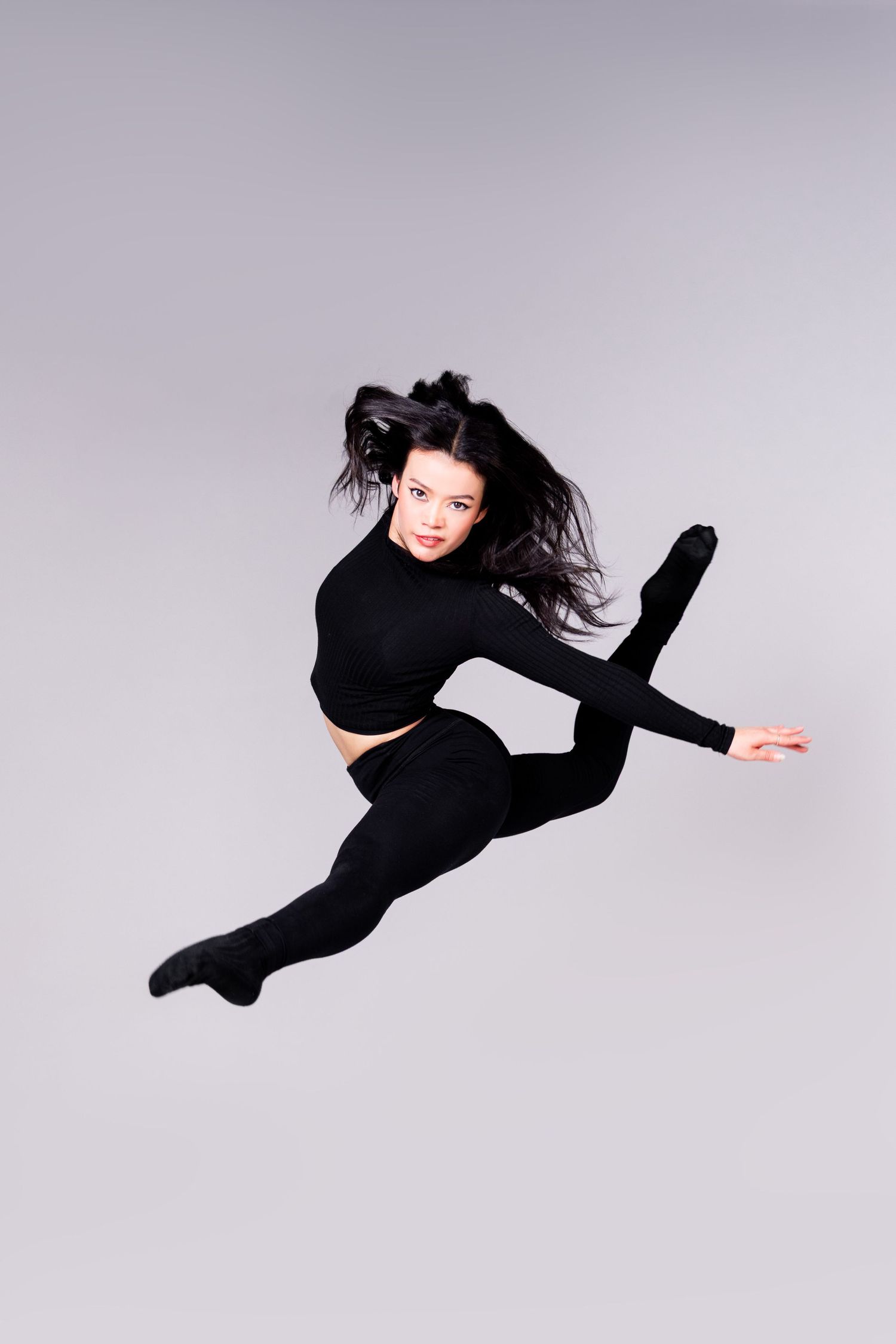 Dancer in black attire performs a dramatic leap with flowing hair against a gray background.