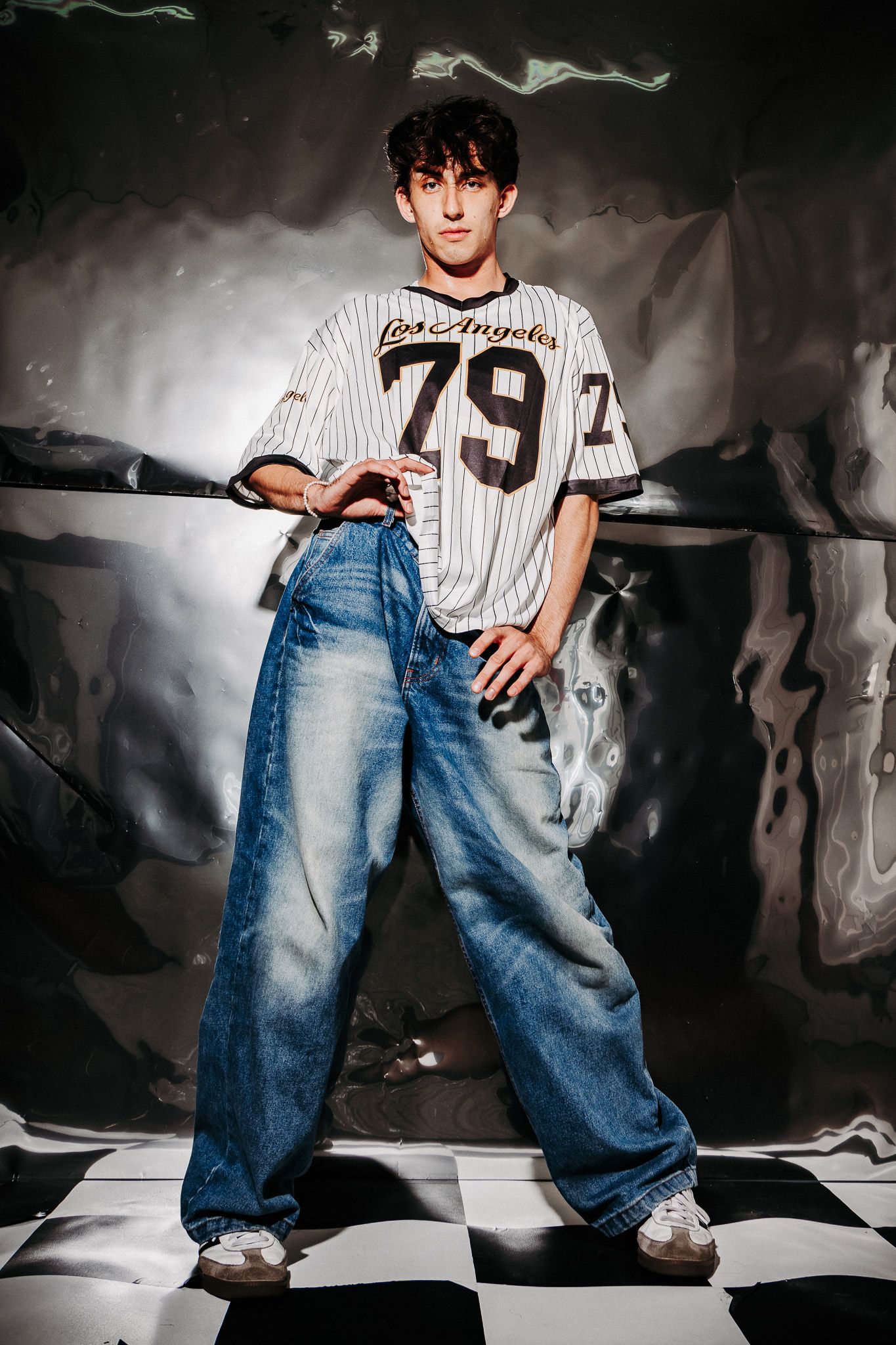 Person wearing baseball jersey number 79 and loose jeans poses on black and white checkered floor against metallic wall.