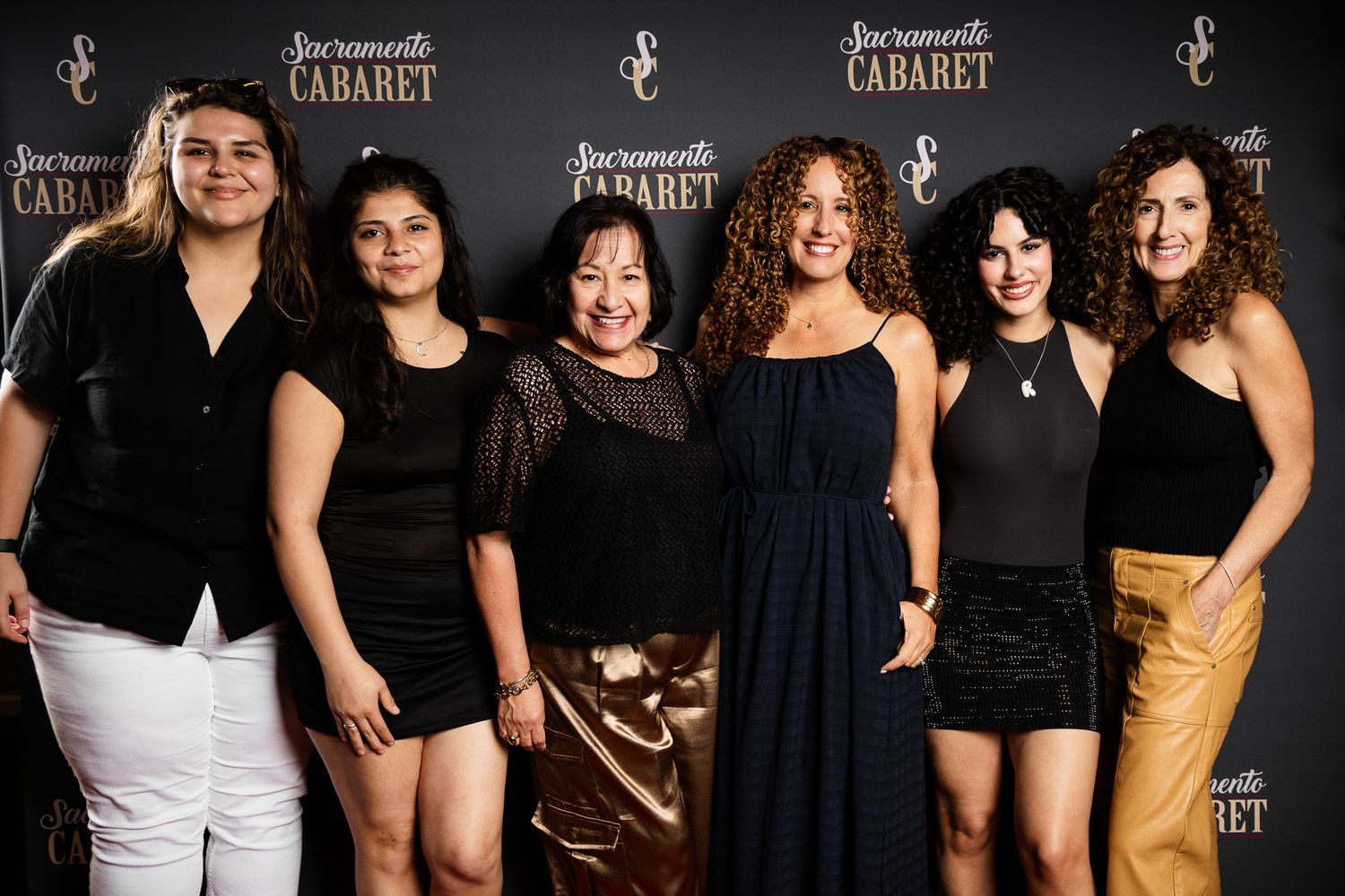 Group of performers pose together in elegant black attire on stage at a cabaret-style performance venue.