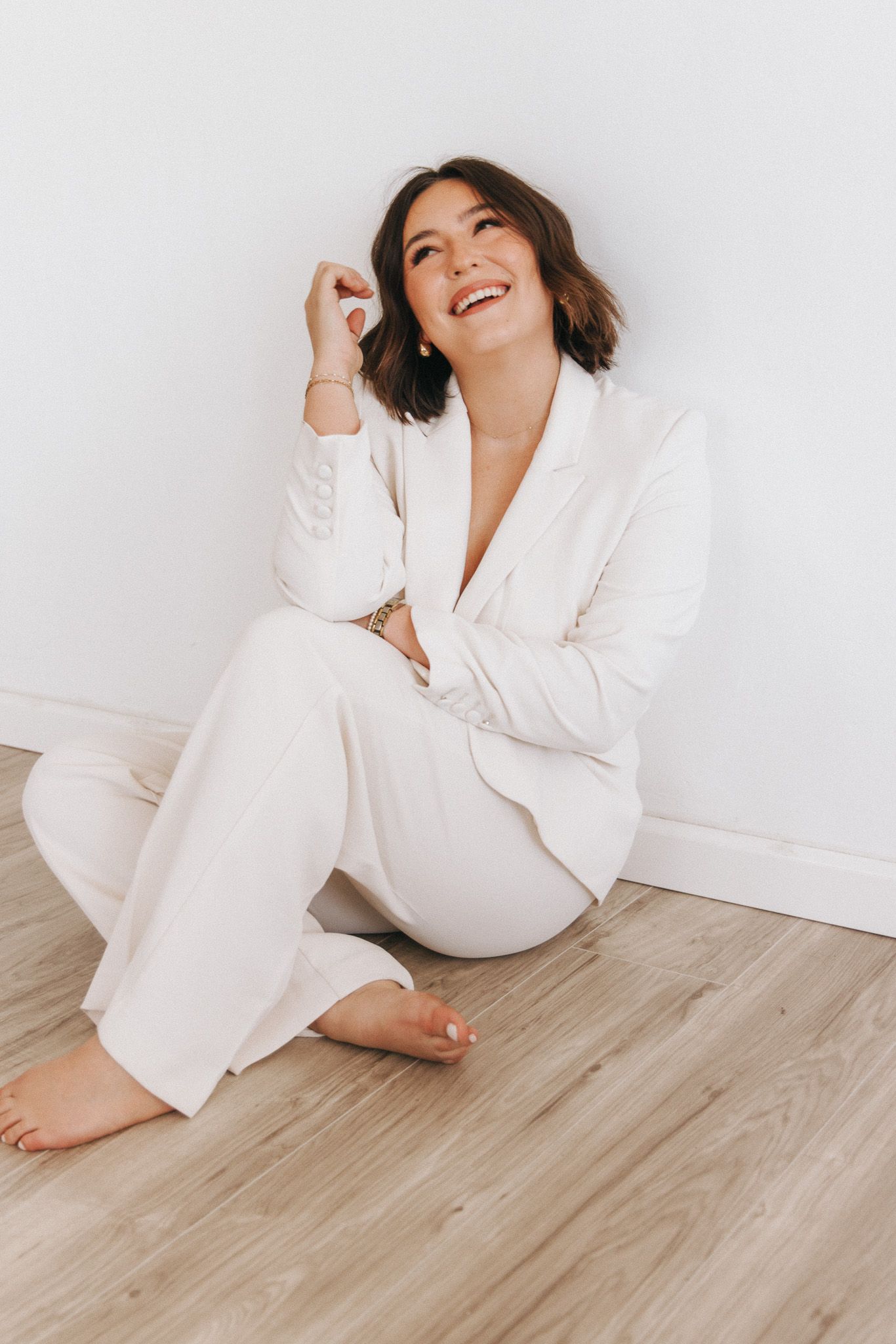 Person wearing white loungewear set sits casually on hardwood floor against white wall while smiling joyfully.
