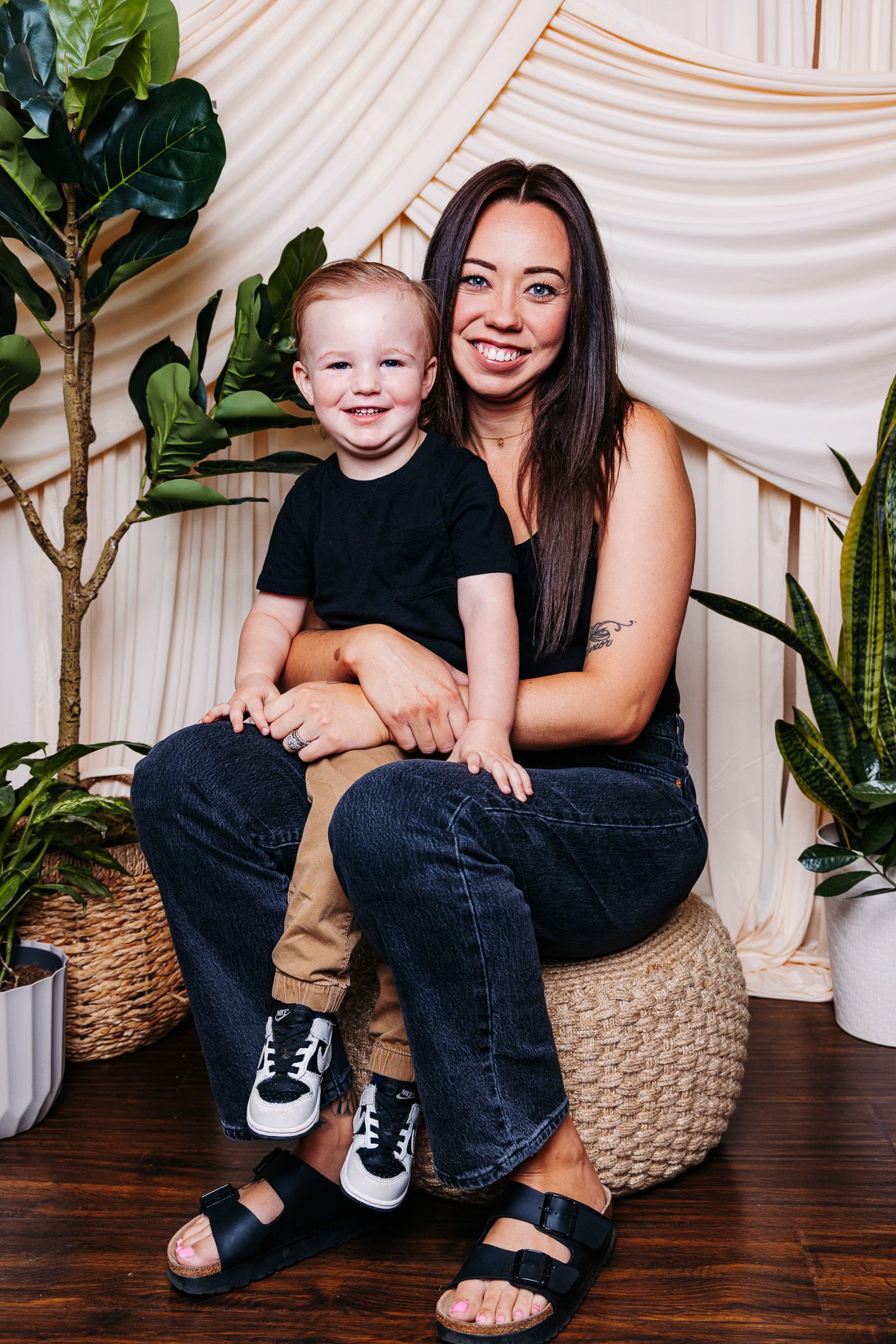 A smiling woman and toddler boy pose together on a woven pouf surrounded by green plants and a cream backdrop.