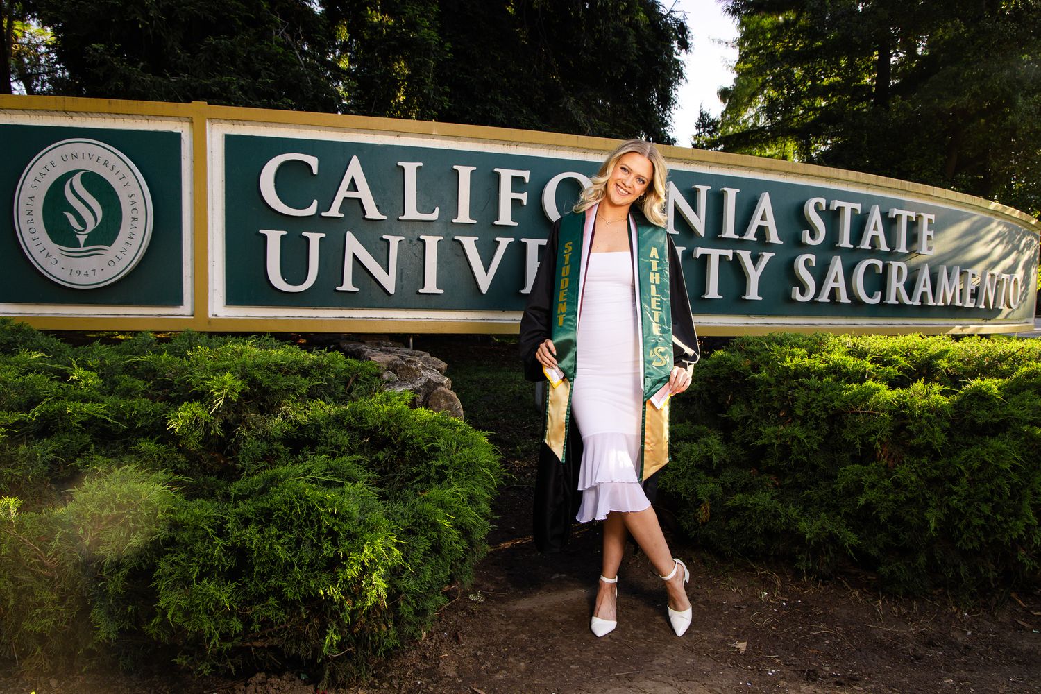 Graduate poses in cap and gown at California State University Sacramento sign outdoors.