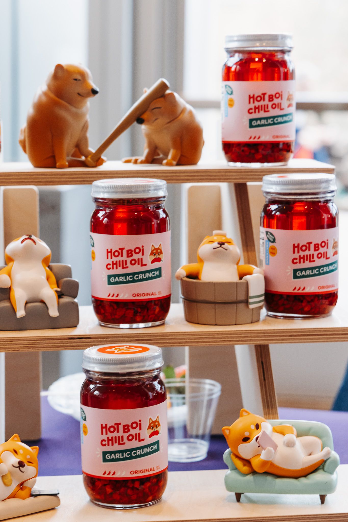 Cute ceramic shiba inu figurines arranged on shelves alongside jars of red chili oil in a whimsical display setup.
