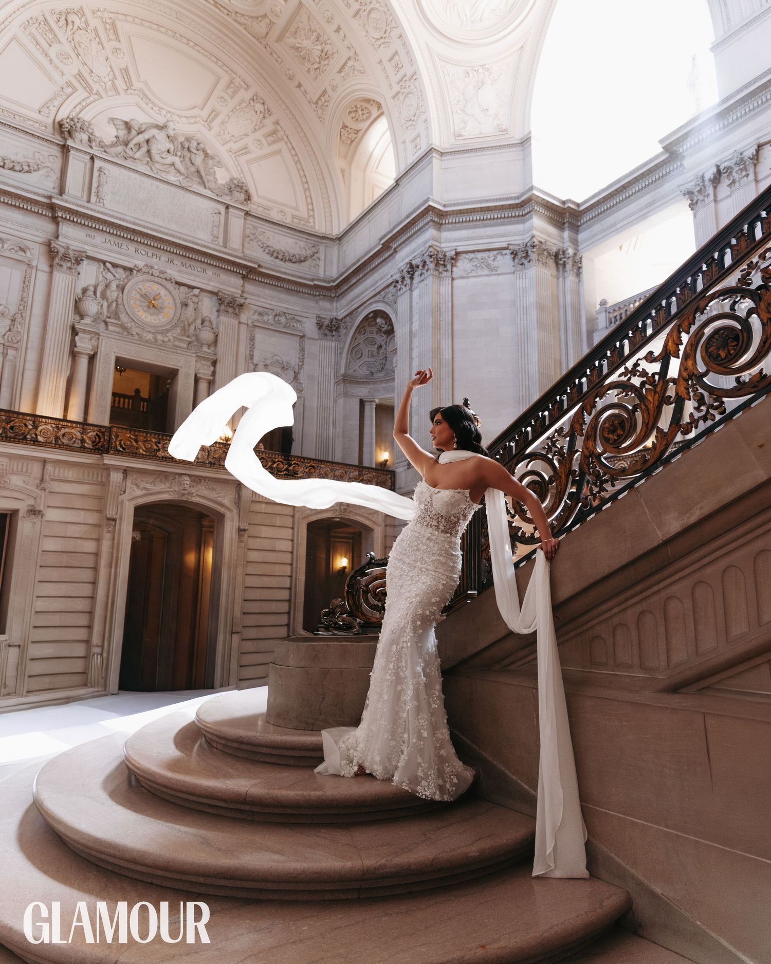 A bride in a white gown poses elegantly on a grand marble staircase with ornate railings in a luxurious palace setting.