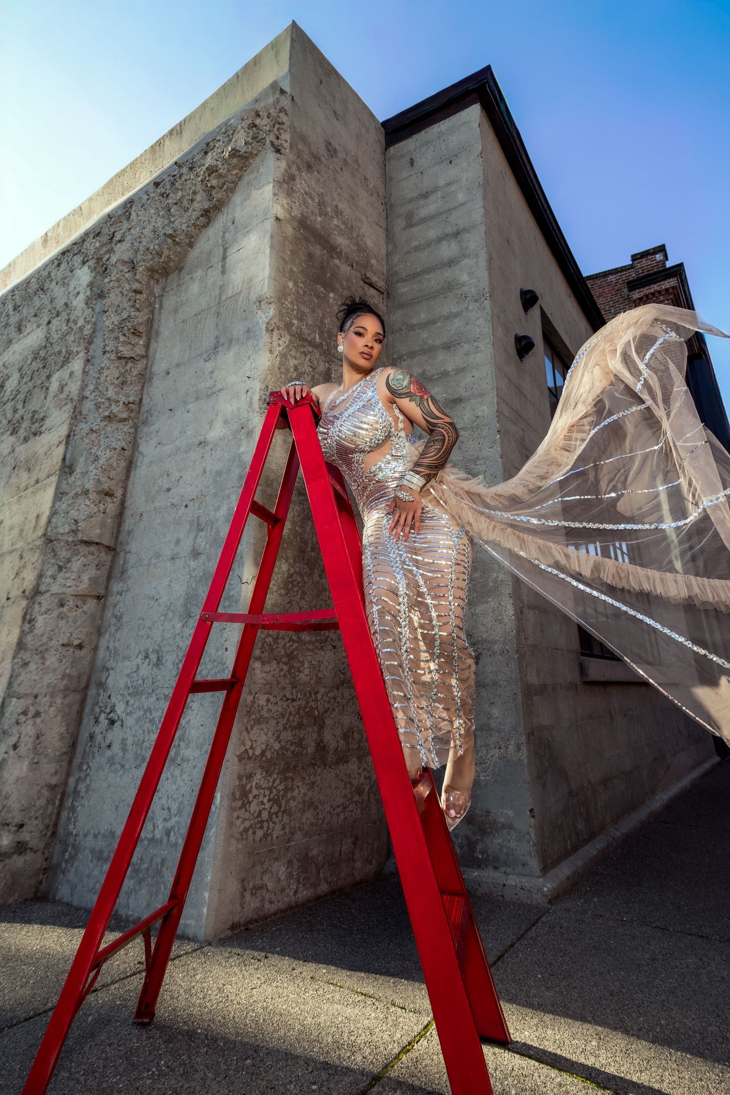 A person in a shimmering gold evening gown poses dramatically on a red ladder against a concrete building exterior.