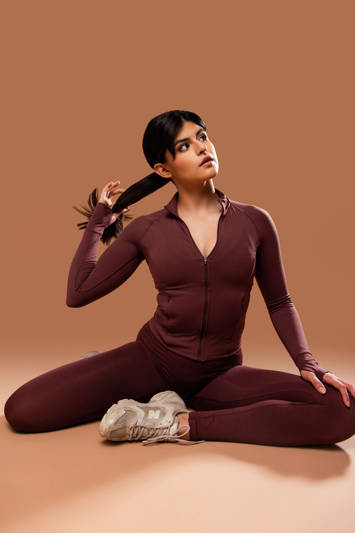 Athletic figure in burgundy workout set poses on brown background while stretching in yoga position.