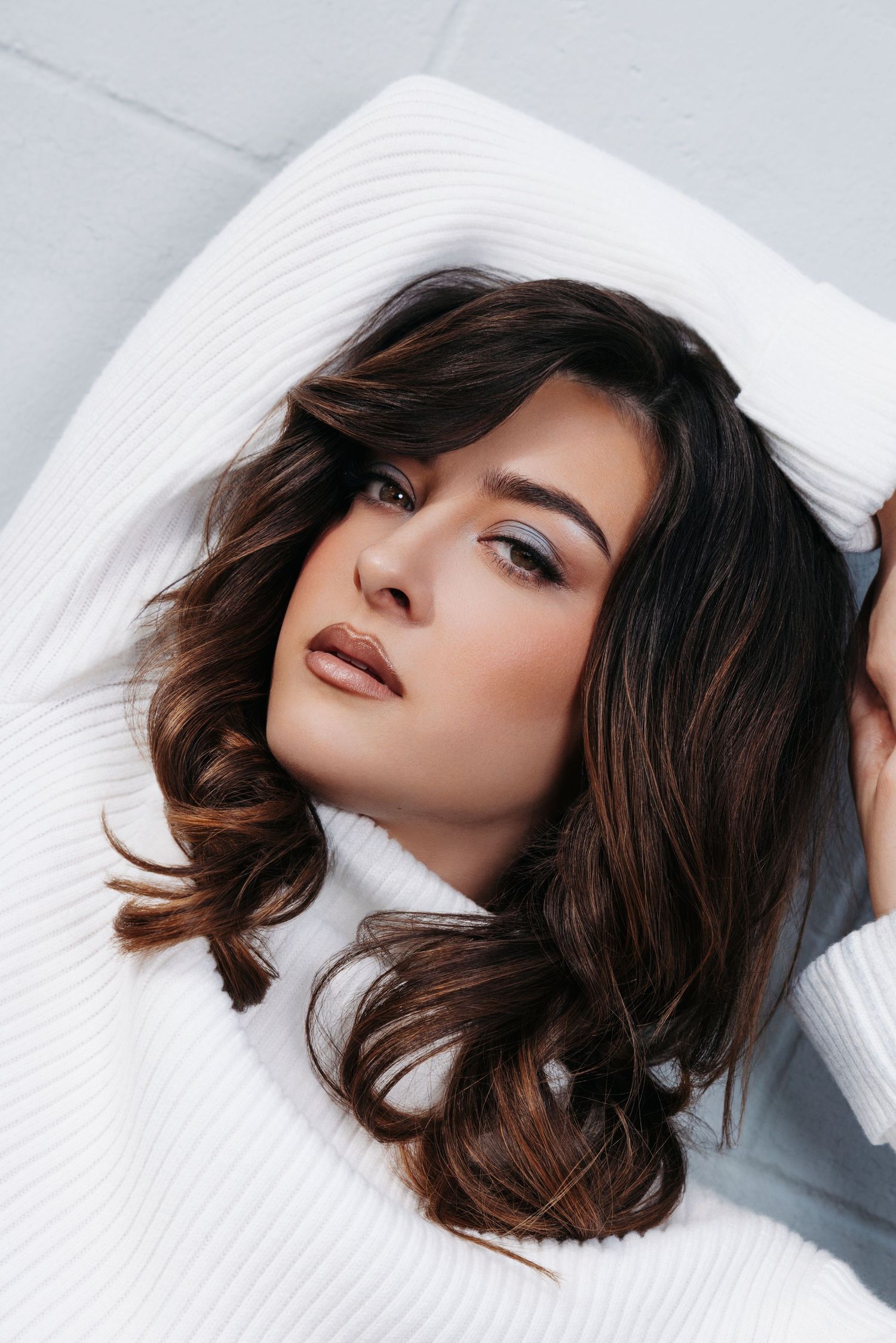 Beauty portrait with dark wavy hair laying on white fabric with dramatic makeup and brown lipstick.