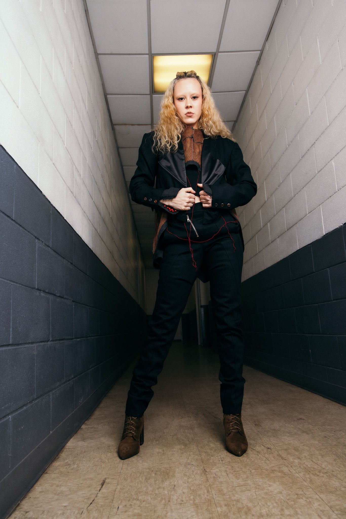 Person in black outfit sits in stark white hallway corner during fashion photoshoot.