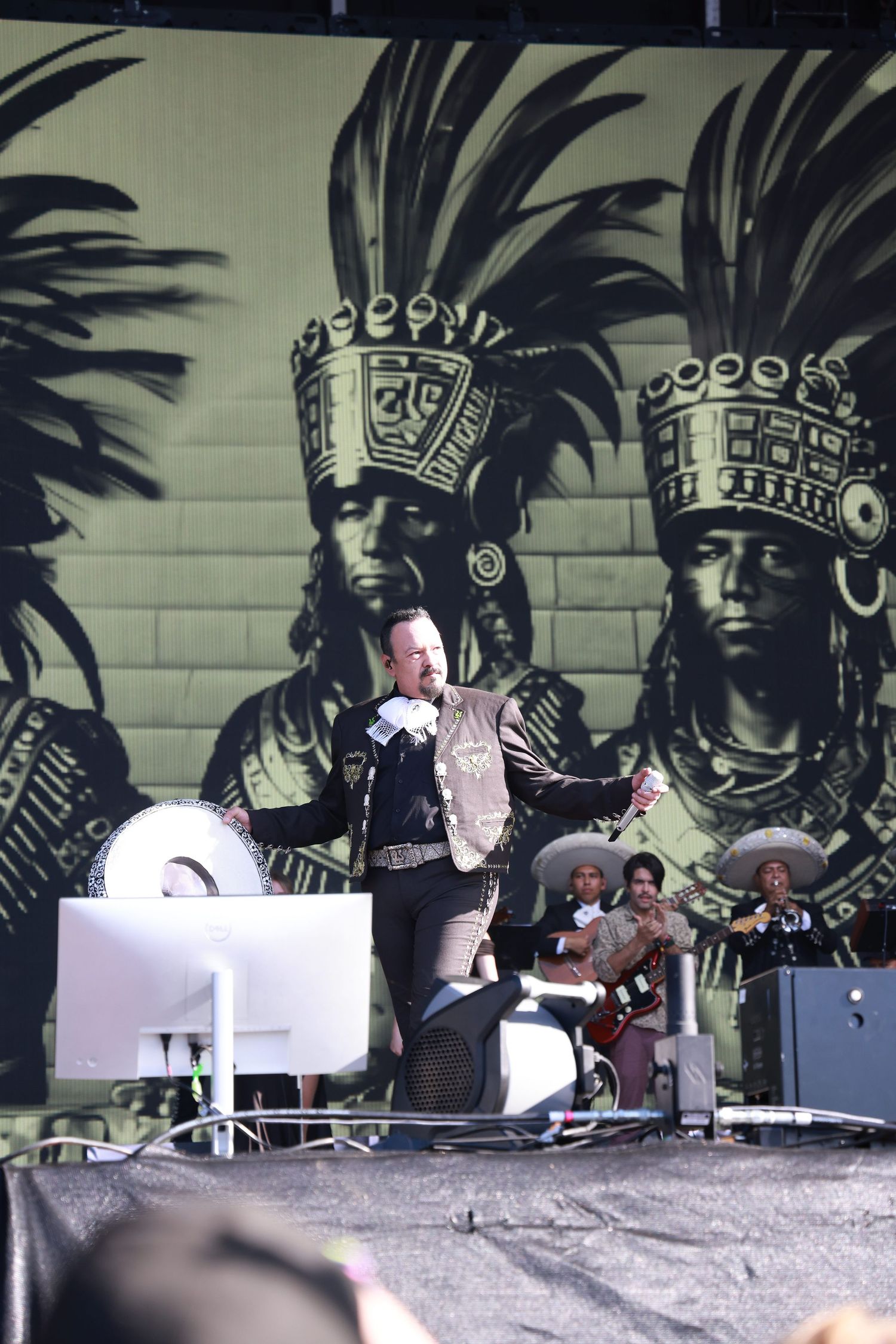 A performer on stage in front of a large mural featuring ancient Aztec or Mayan warrior designs.