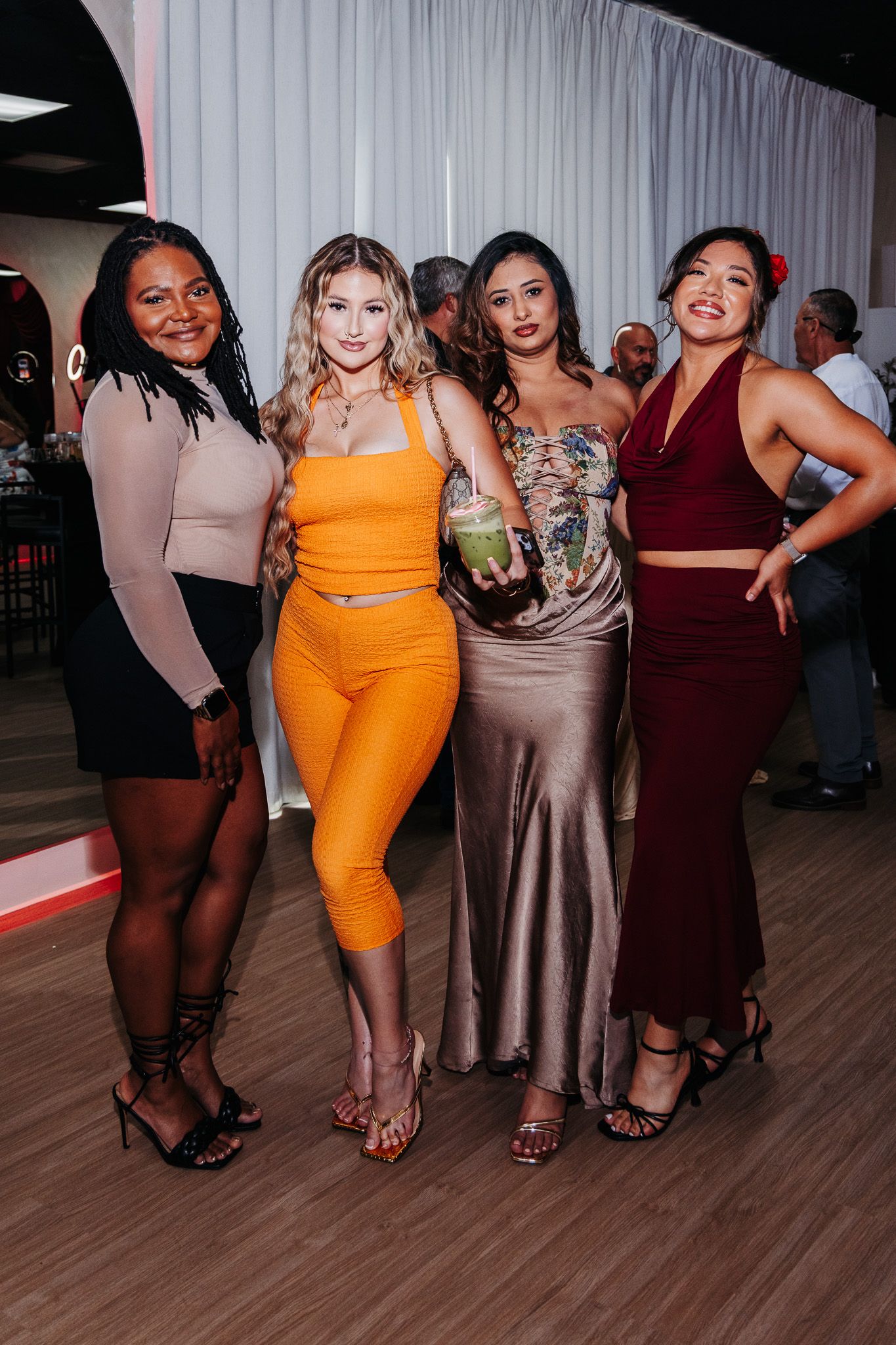 Group of women in formal dresses showing off their footwear and outfits at an event on wooden flooring.