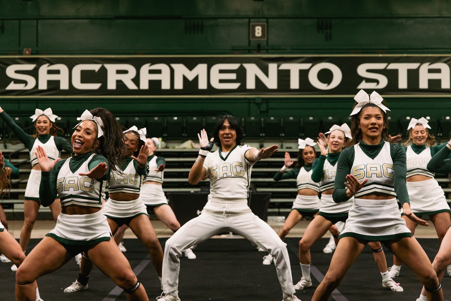 Sacramento State cheerleaders performing energetic routine in green and white uniforms on indoor court.